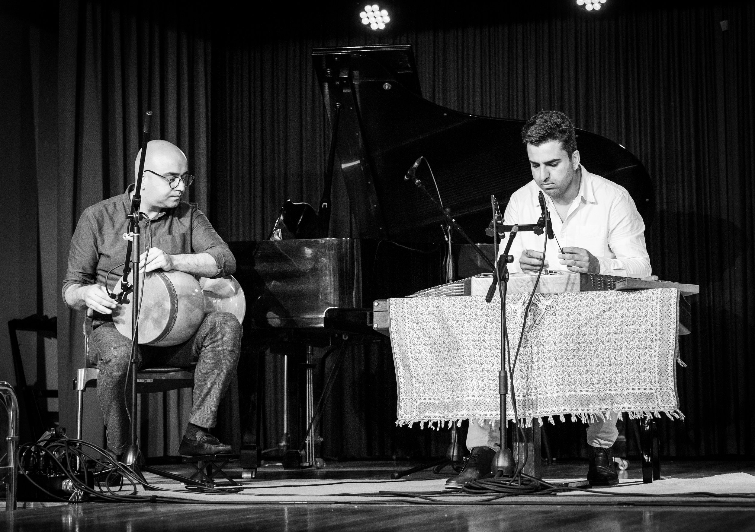 Two musicians on stage, one playing a drum and the other playing a string instrument, with a piano in the background.