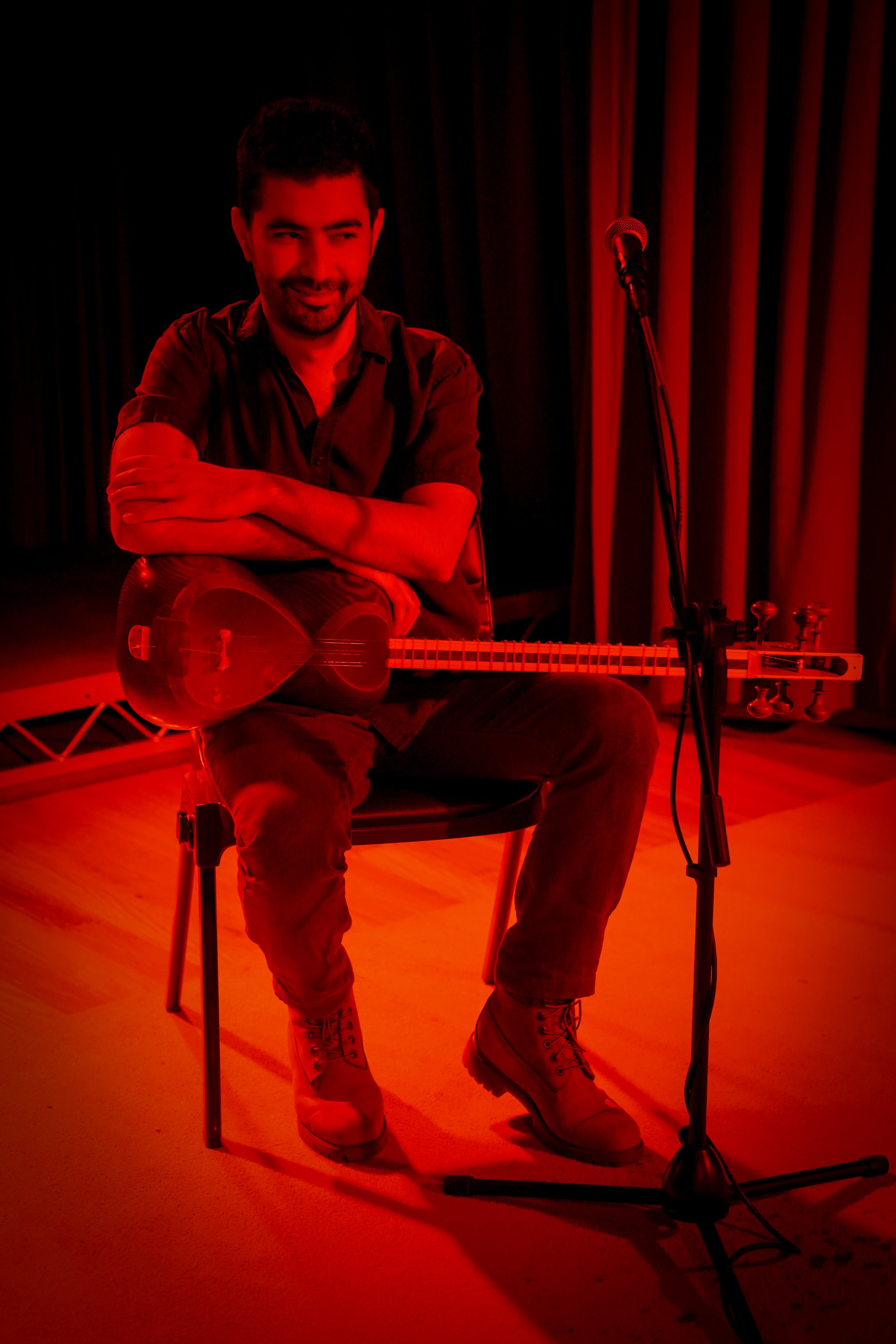 Person sitting on a chair holding a stringed musical instrument with a microphone on a stand in front, under red lighting.