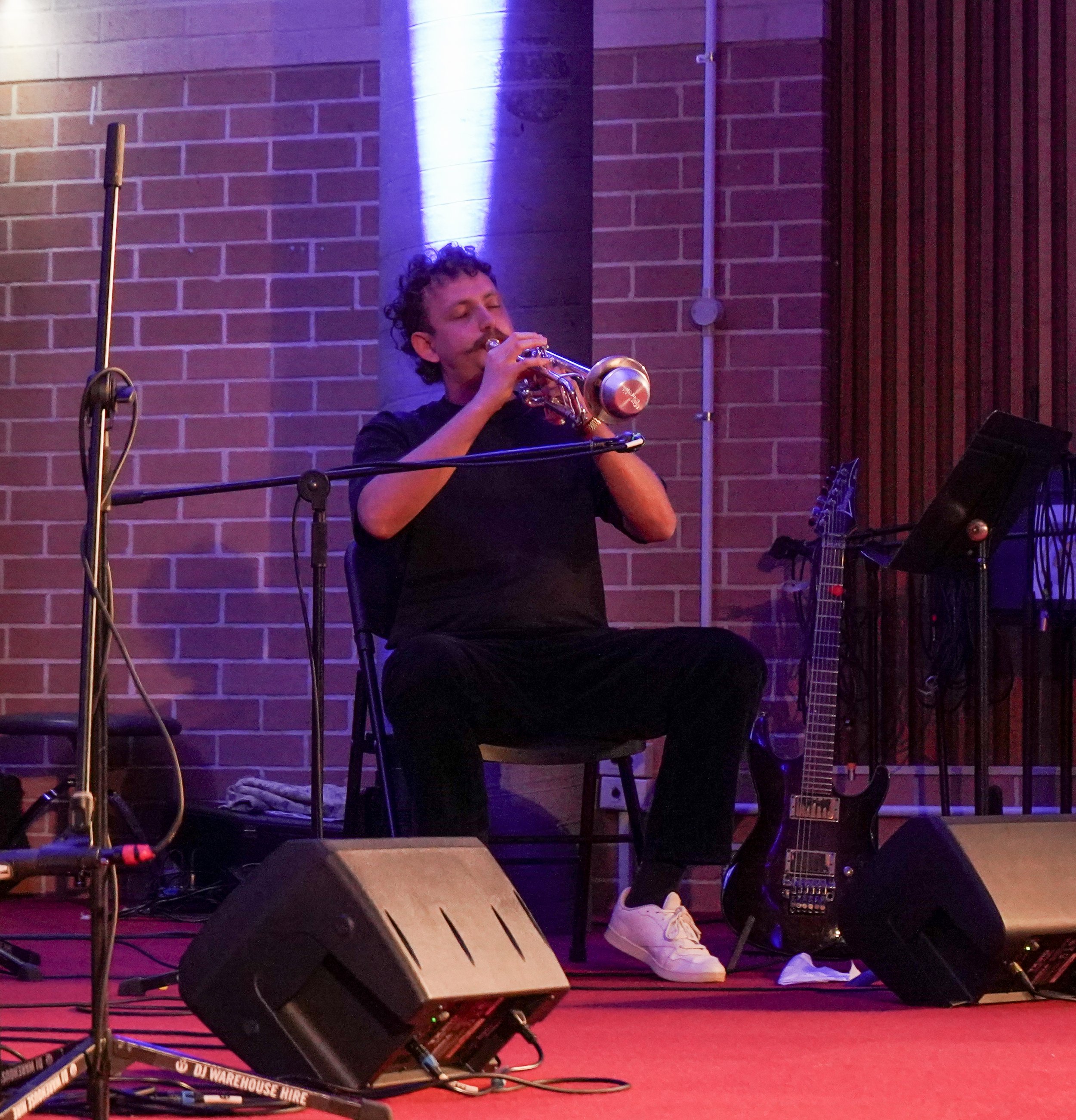 A musician seated on stage playing a trumpet, surrounded by microphones and musical equipment, with a guitar resting against a wall in the background.