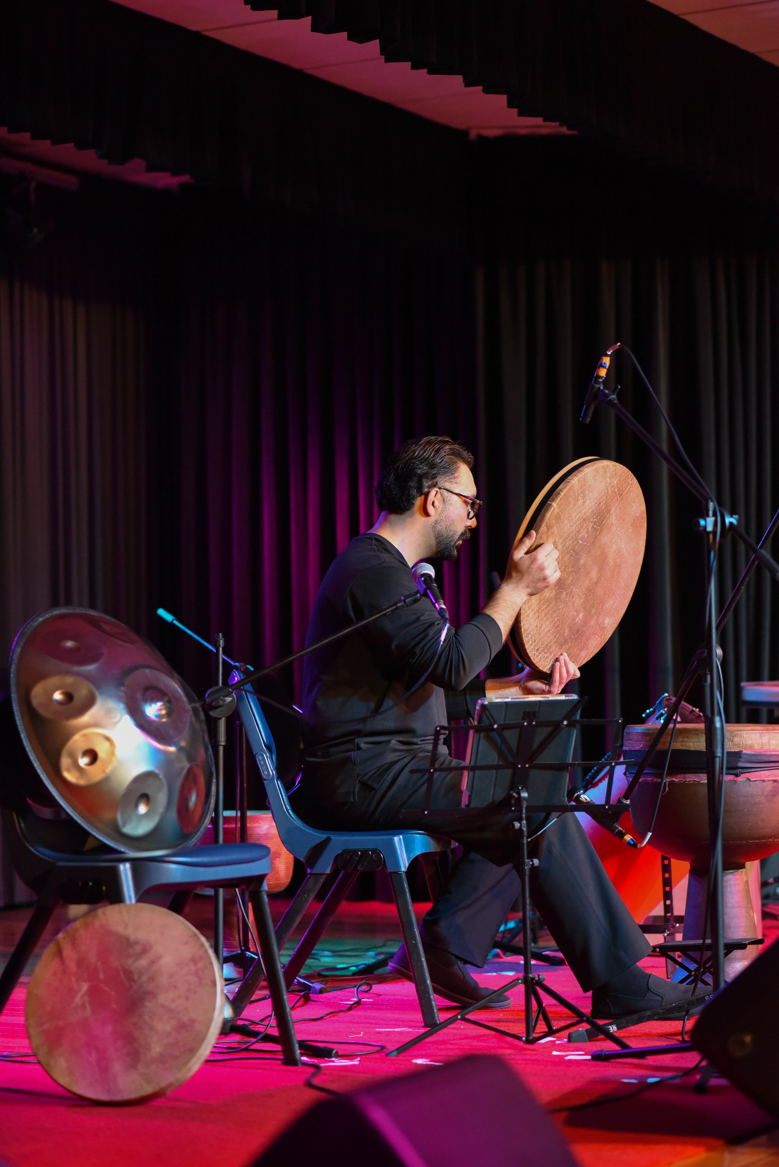 Musician playing a traditional drum with microphones on stage, additional drums nearby, seated in front of a dark curtain.
