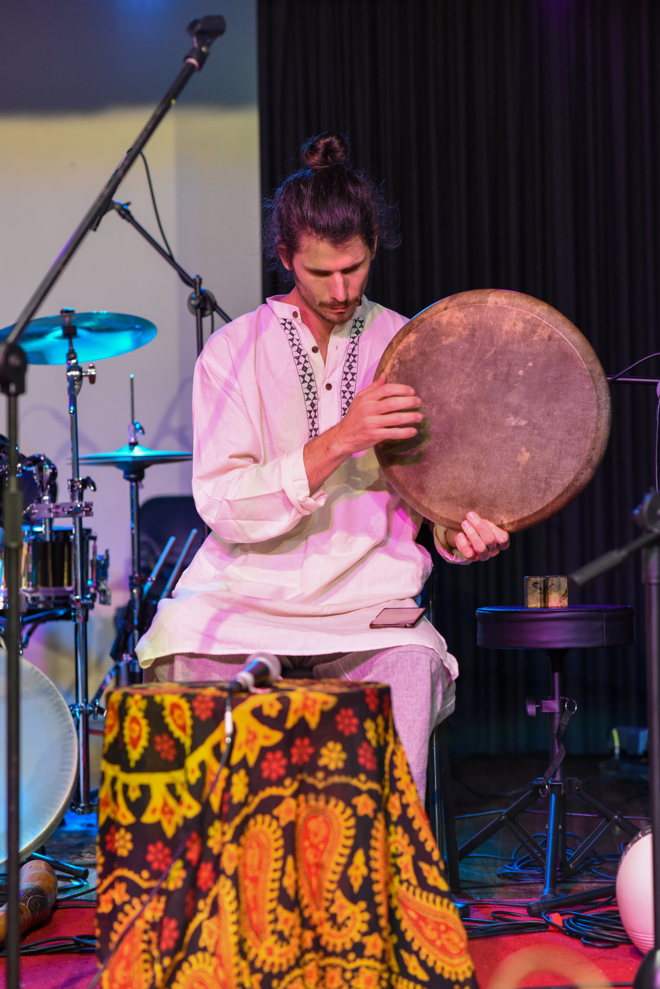 Musician playing a traditional hand drum on stage, wearing a white embroidered shirt, with other musical instruments like a drum set and microphones in the background.