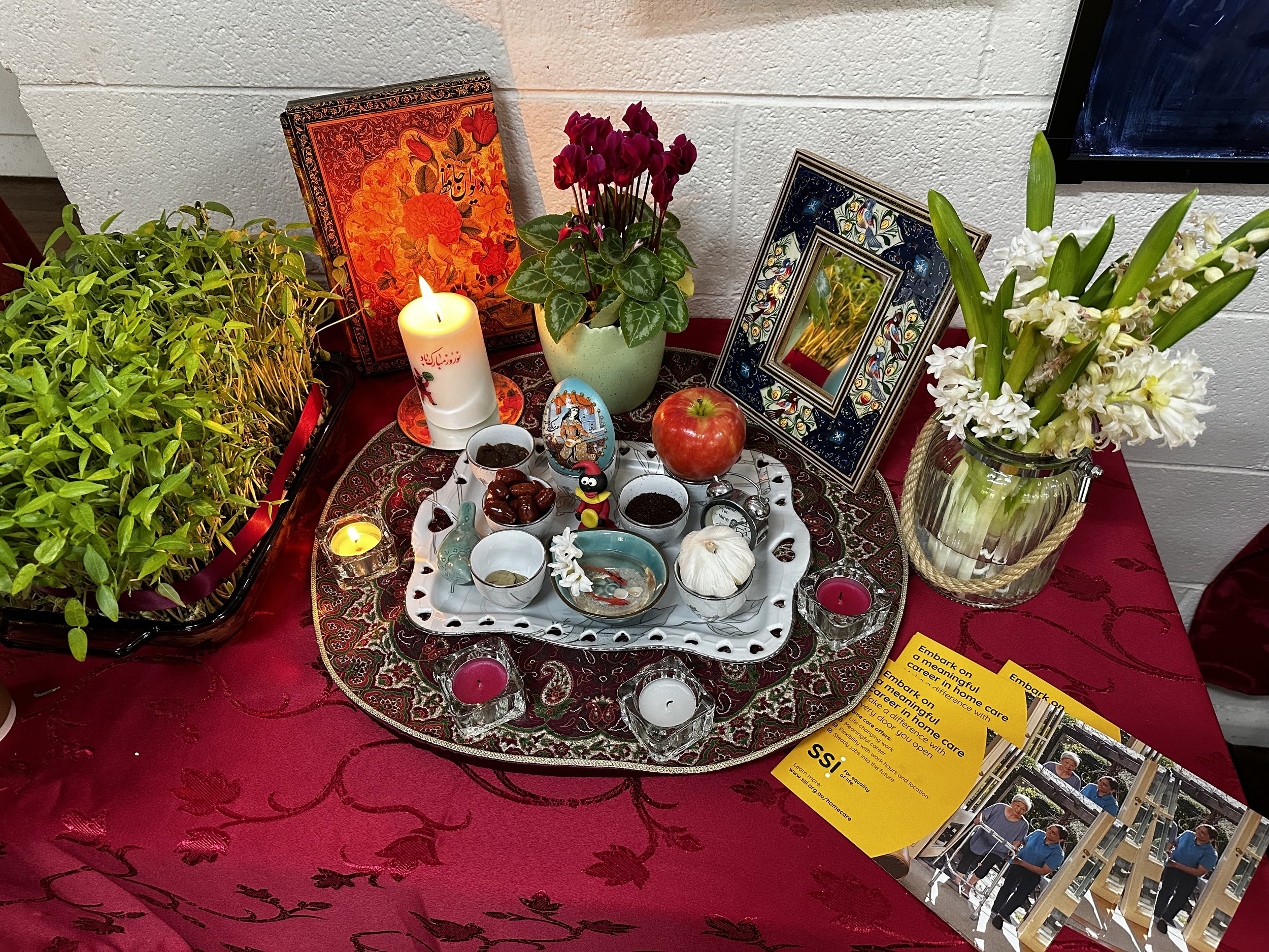 Persian New Year Haft-Seen table with traditional items like a candle, mirror, apple, garlic, sweets, and plants on a red cloth.