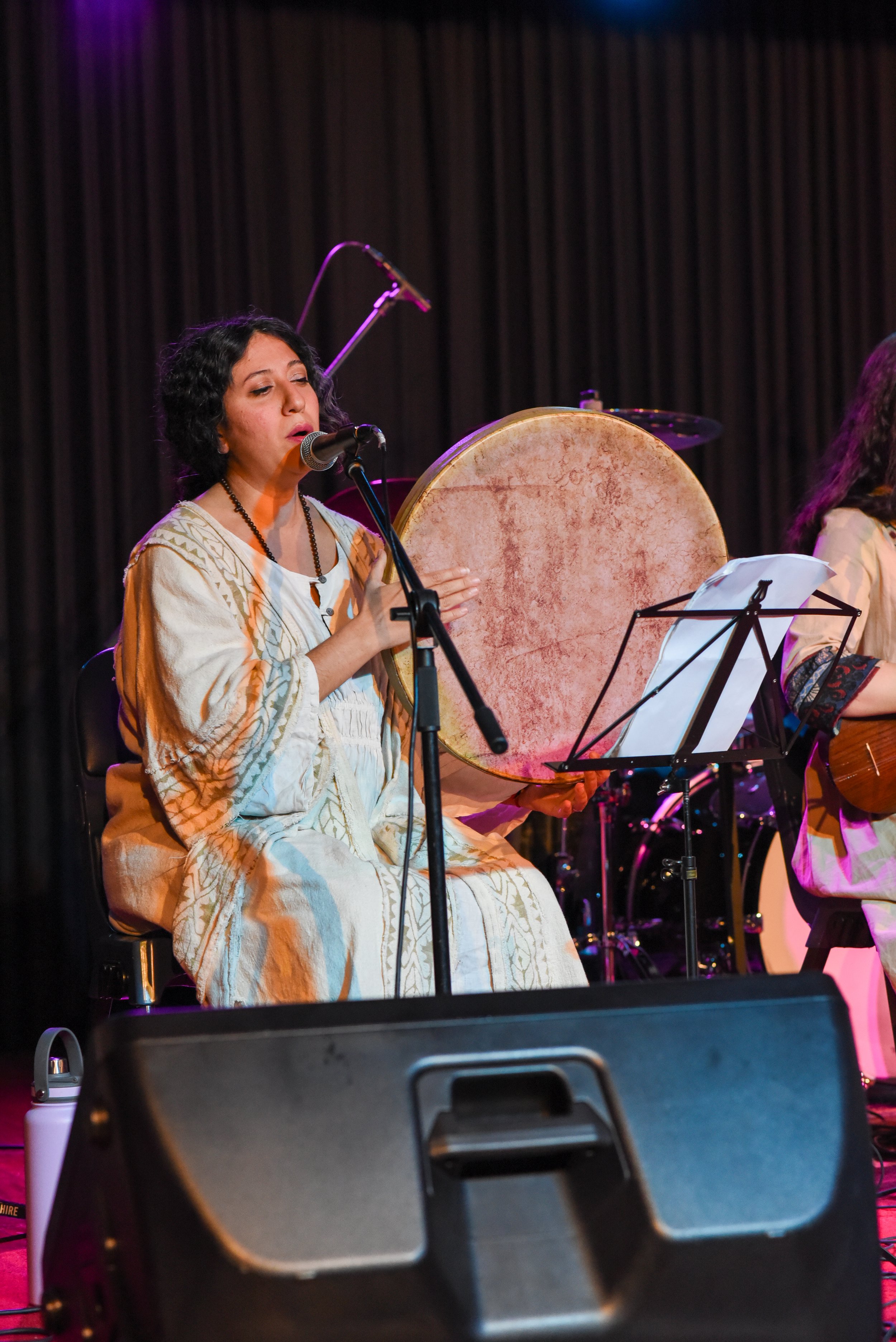 A musician in a white traditional outfit singing into a microphone and playing a large frame drum on a stage, with musical notes on a stand beside them.