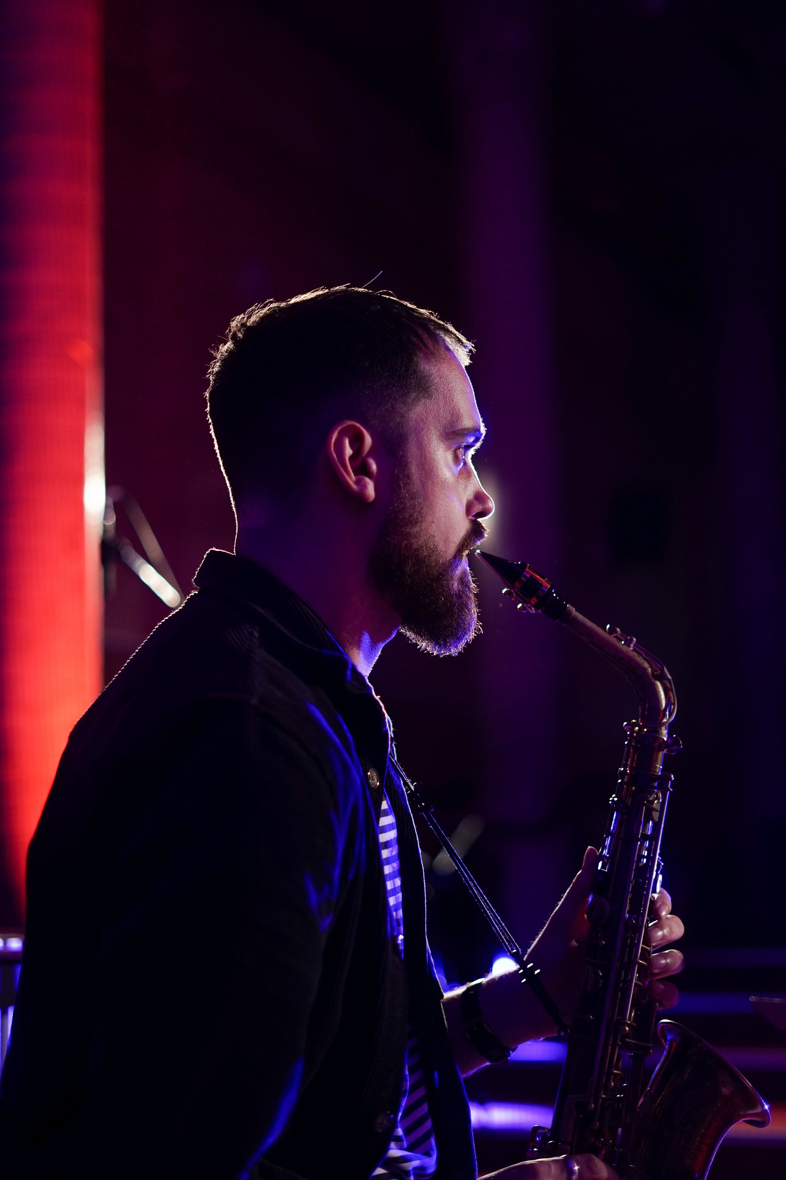 Side view of a man playing a saxophone on stage with colorful lighting.