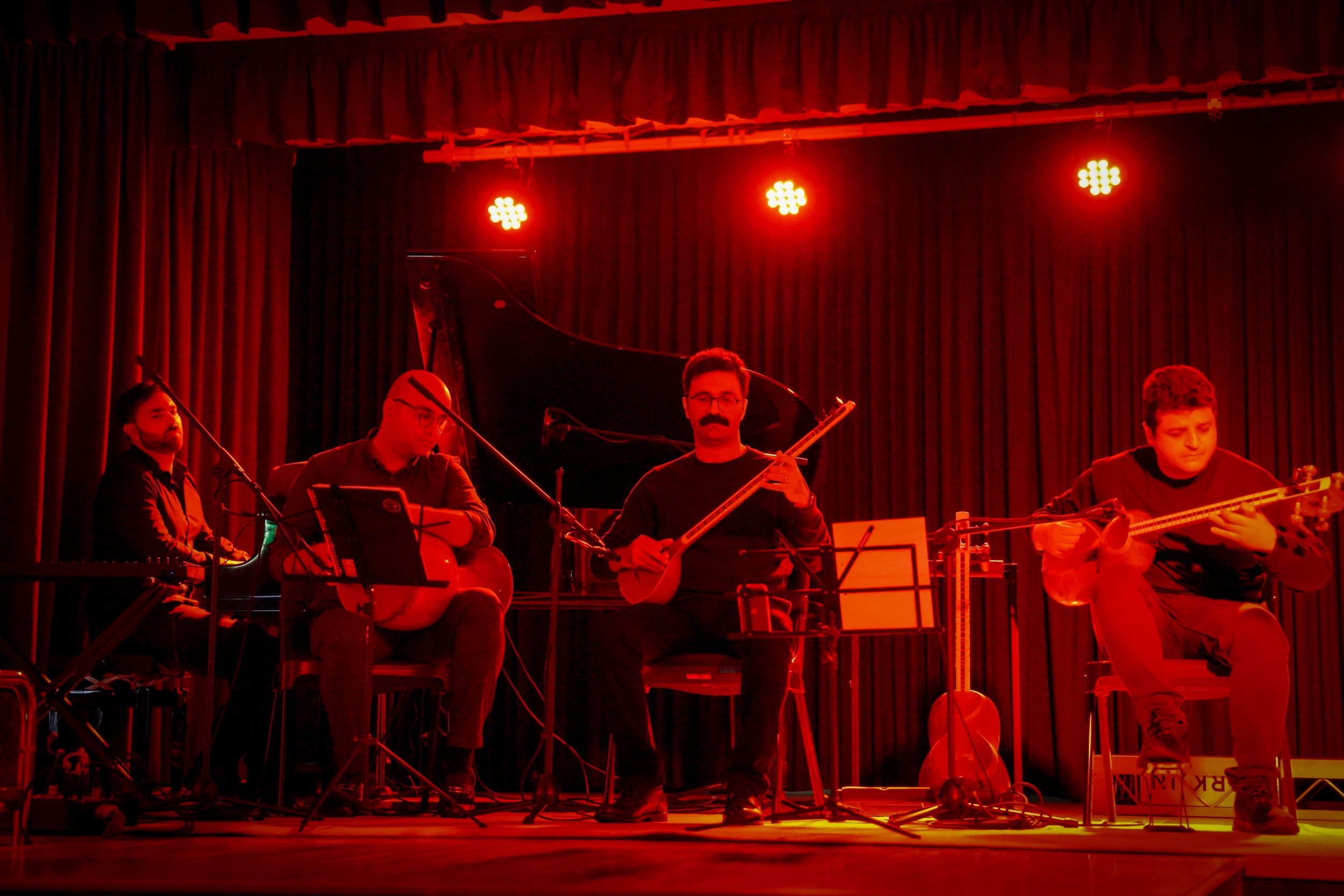 Musicians performing on stage with string instruments and piano under red lighting.