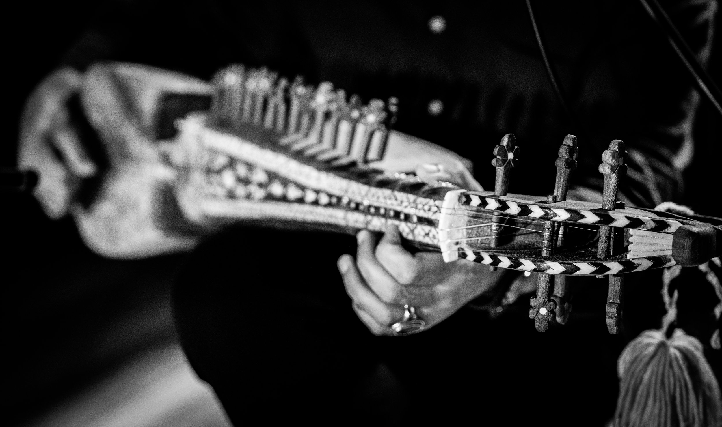 Black and white image of a person playing a traditional string instrument, possibly a lute or similar, with intricate tuning pegs and hand-carved details.