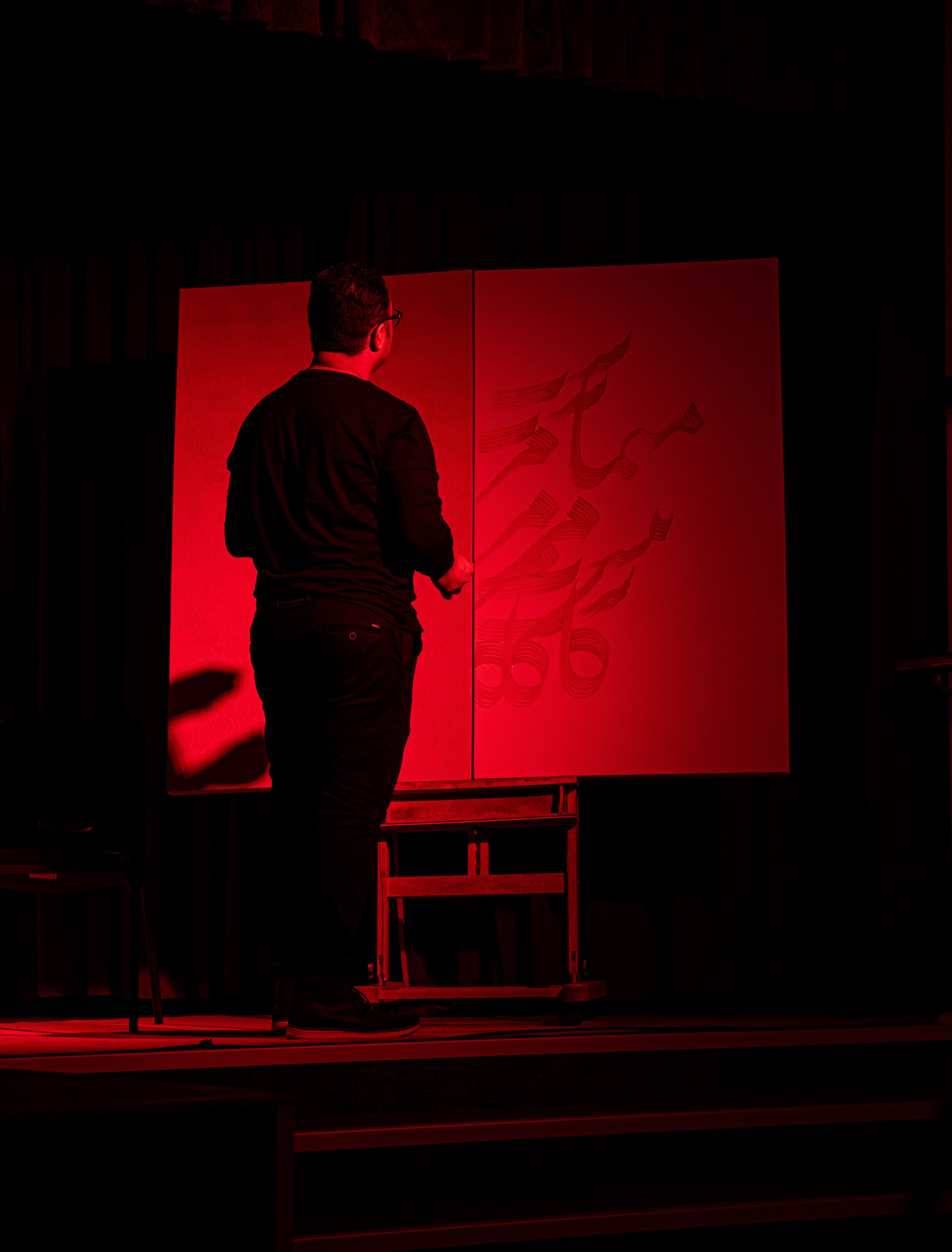 Person painting on a large canvas under red stage lighting, creating abstract calligraphy or design.