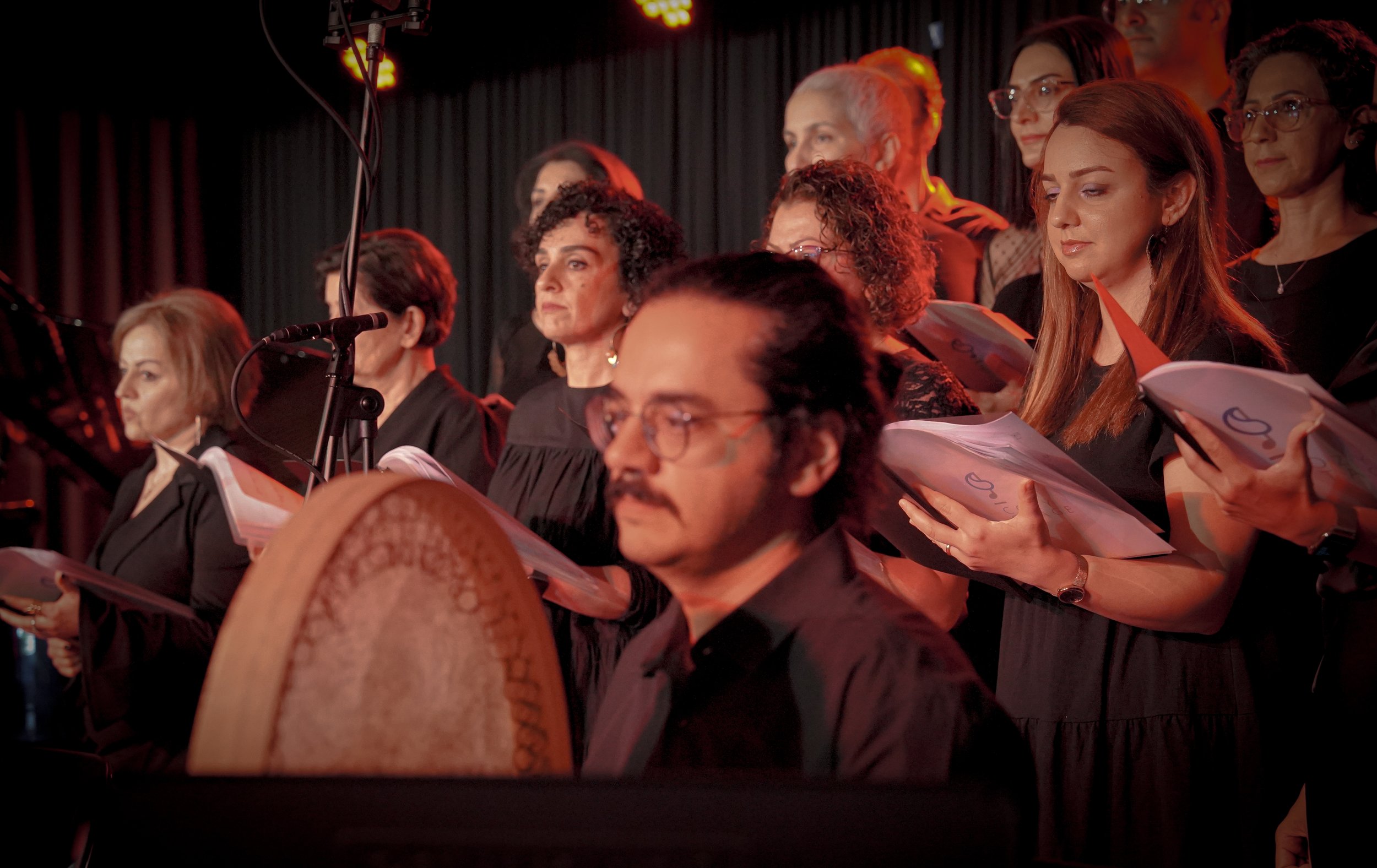 A choir group performing on stage with a percussionist in the foreground, holding a large frame drum. The choir members are holding sheet music and are illuminated by stage lighting. The background is a dark curtain.