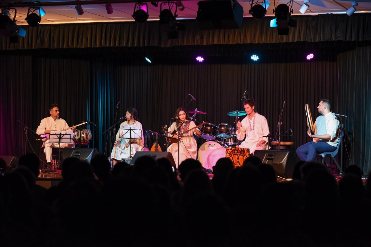 A musical band performing on stage with traditional instruments and a backdrop of dim lighting. Five musicians are seated, wearing traditional attire, playing various instruments including drums and string instruments, under colorful stage lights.