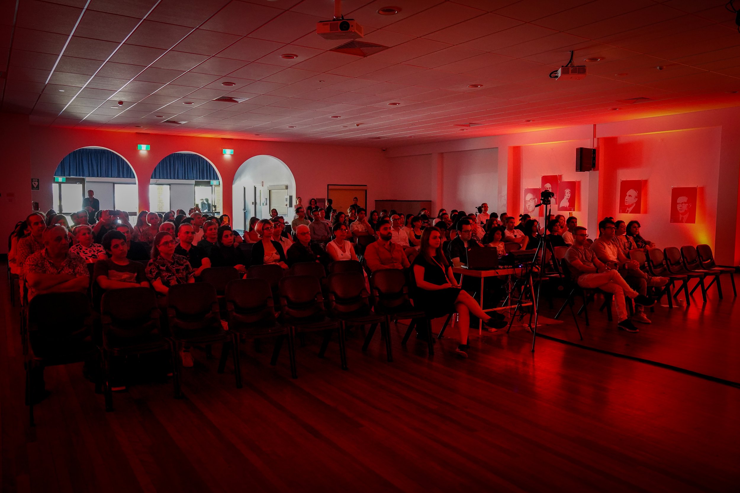 Audience seated in a dimly lit conference room with red lighting, arches, and portraits on the wall.