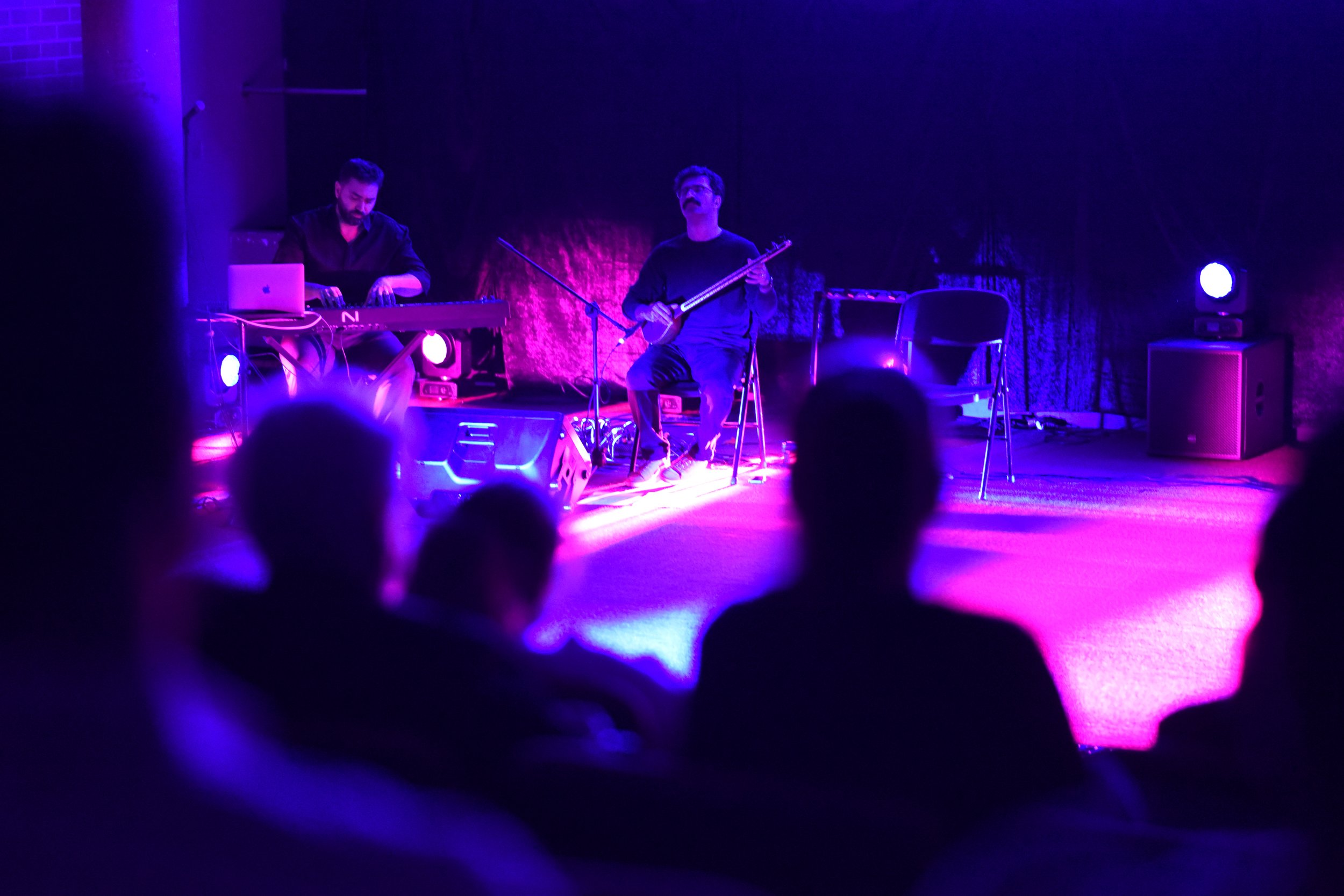 Musicians performing on stage with purple lighting, featuring a keyboard player and a guitar player, viewed from an audience.