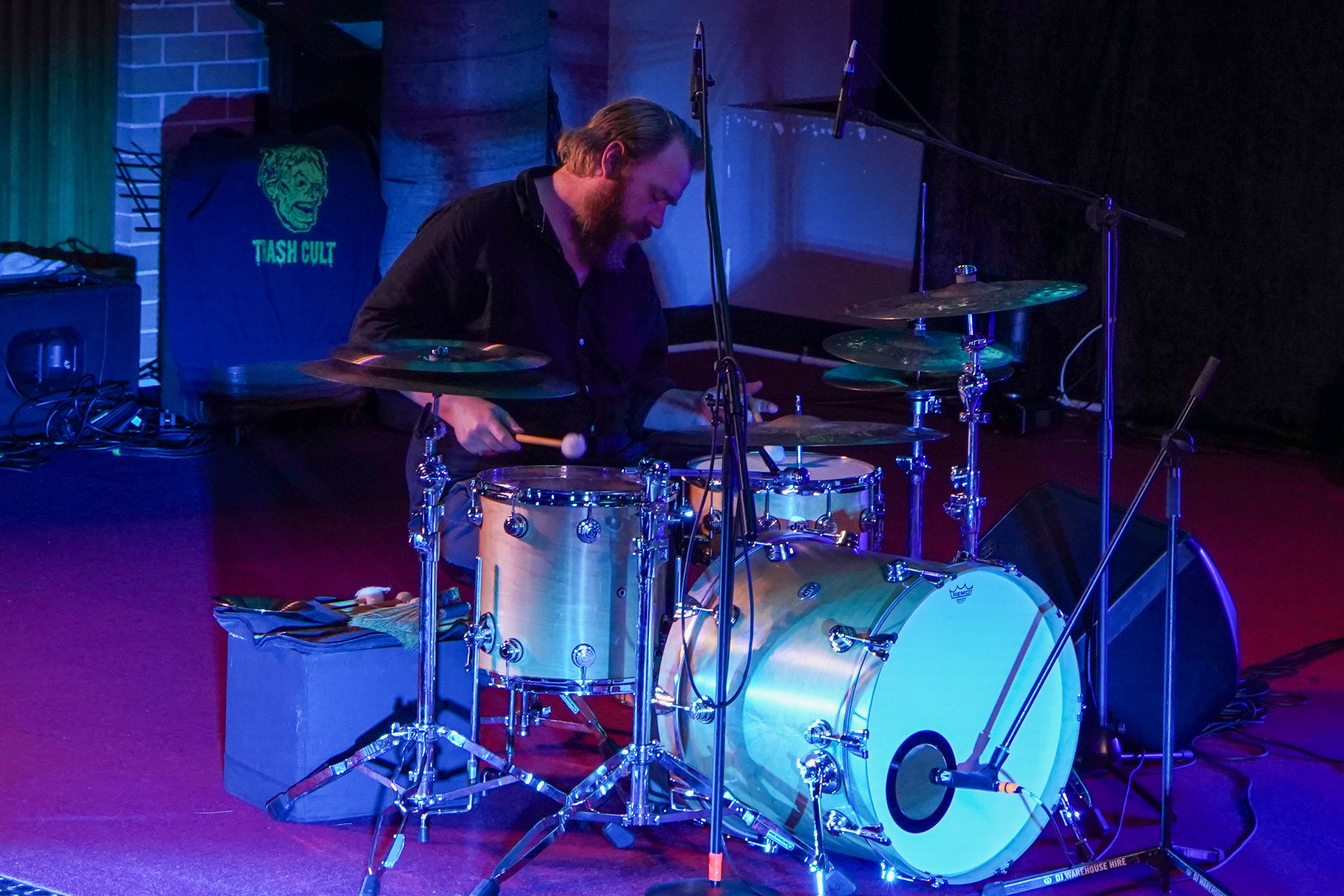 Drummer playing a drum set on stage with microphones and sound equipment around.