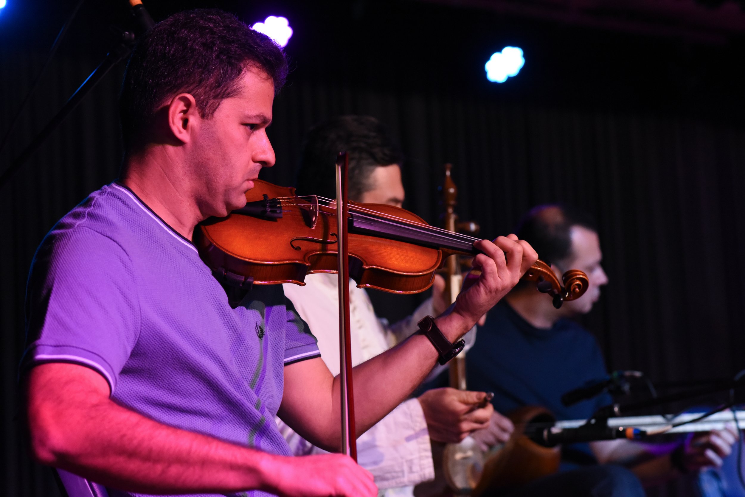 Musician playing violin on stage with others in background.