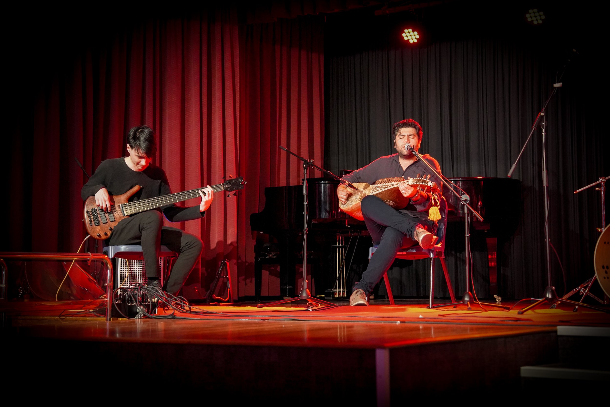 Two musicians performing on stage, one playing an electric guitar and the other playing a traditional string instrument while singing, with red lighting and curtains in the background.