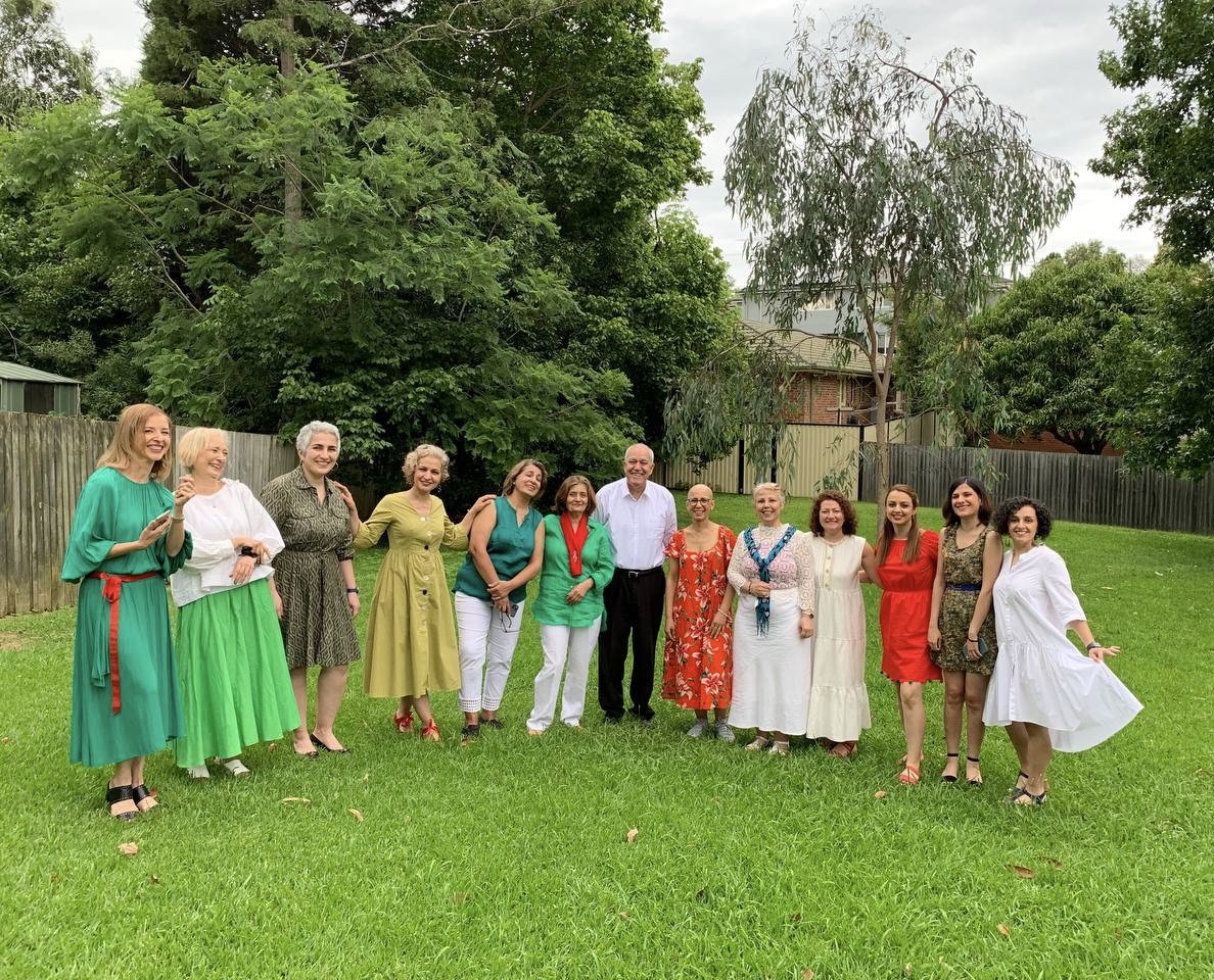 A group of people posing outdoors on a grassy area with trees and a fence in the background. They are dressed in a variety of colorful outfits, including green, red, and white clothing. The atmosphere appears relaxed and cheerful.
