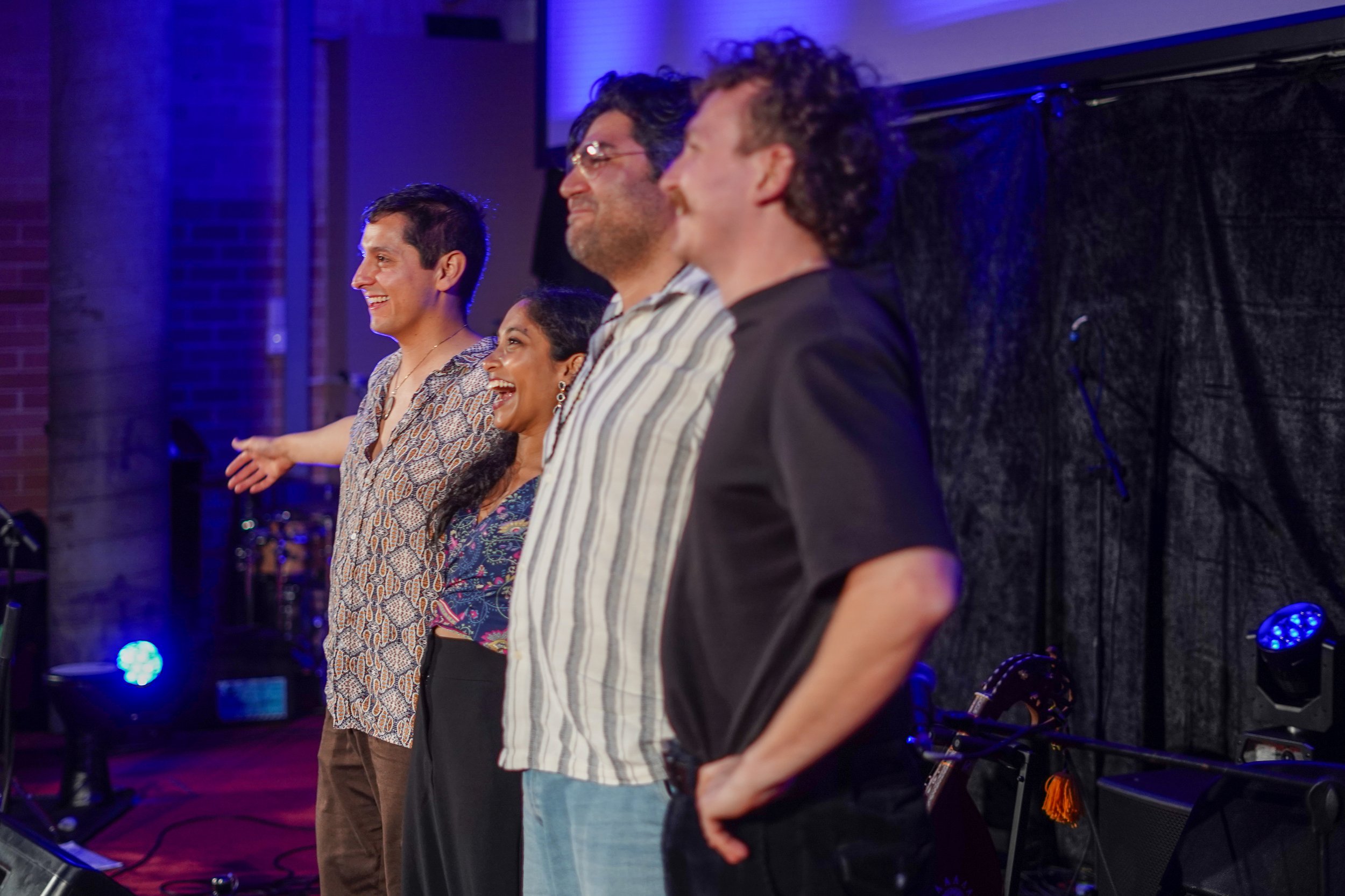 Four people standing in a line on a stage, smiling and looking towards the audience, with stage lighting and musical instruments visible in the background.