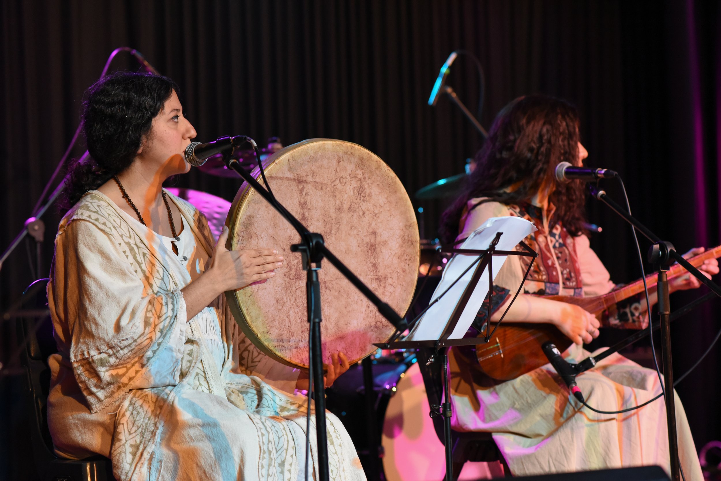 Two women dressed in traditional attire performing on stage; one playing a large frame drum and singing into a microphone, the other playing a stringed instrument.