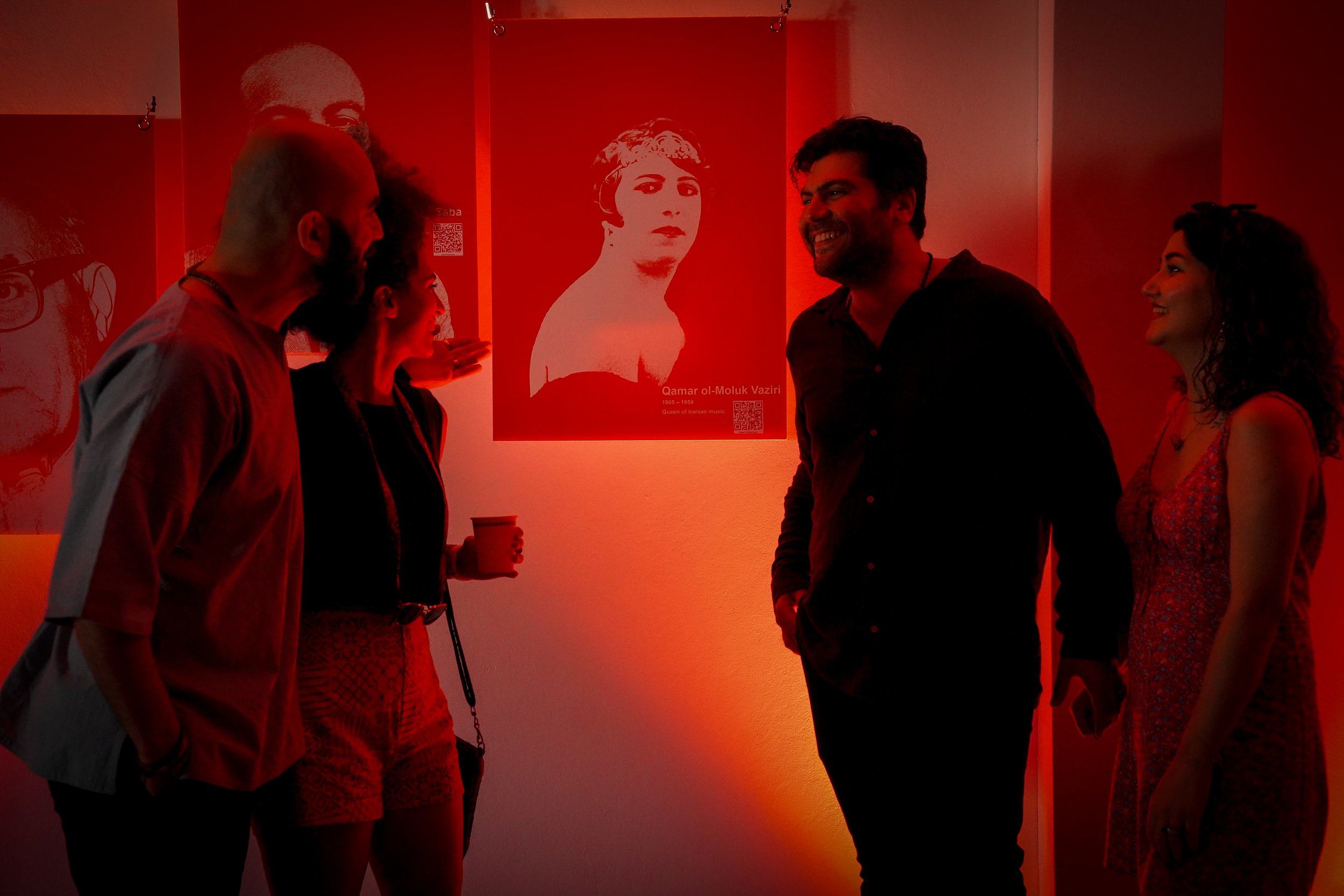 Four people in a dimly lit room with red lighting, looking and smiling at a poster of a historic figure, labeled 'Qamar ol-Moluk Vaziri.'