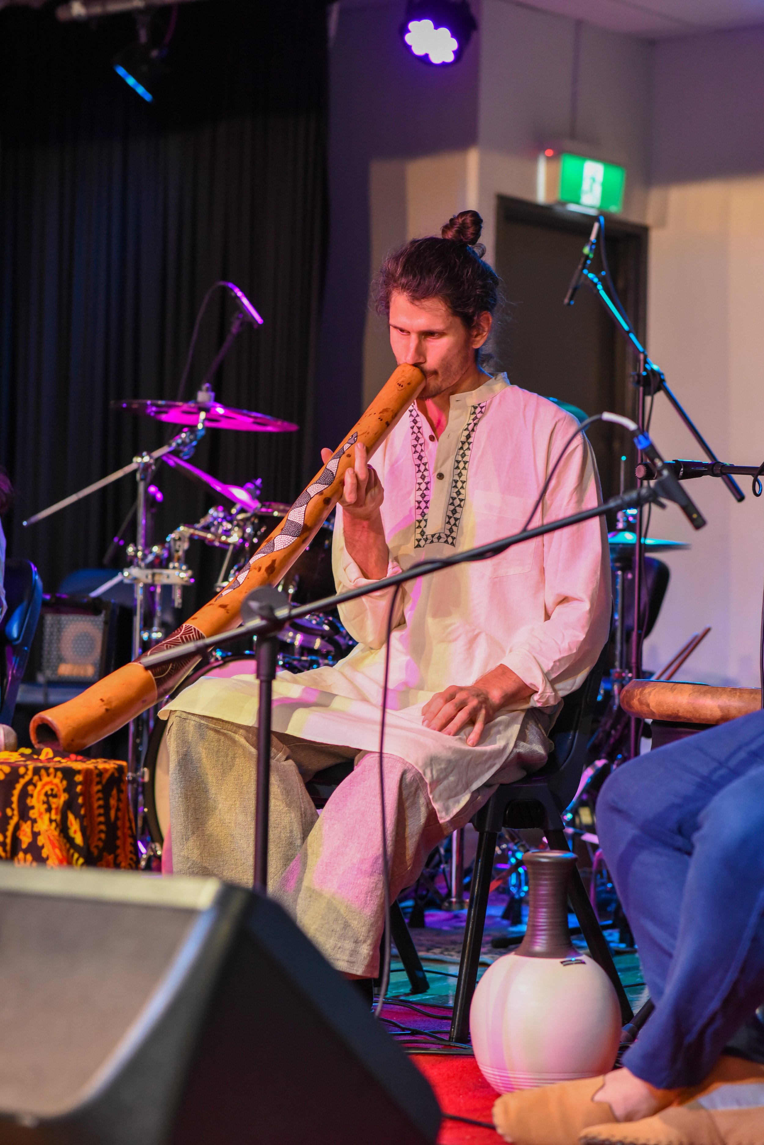 Musician playing a didgeridoo on stage, surrounded by drums and musical equipment, dressed in a white tunic and light pants.