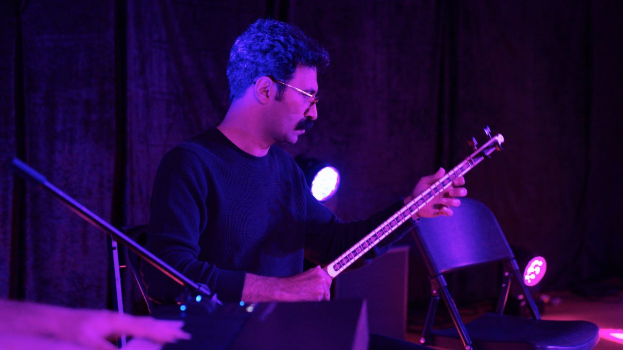 Person playing a tar instrument on stage with colorful lighting and a microphone stand.