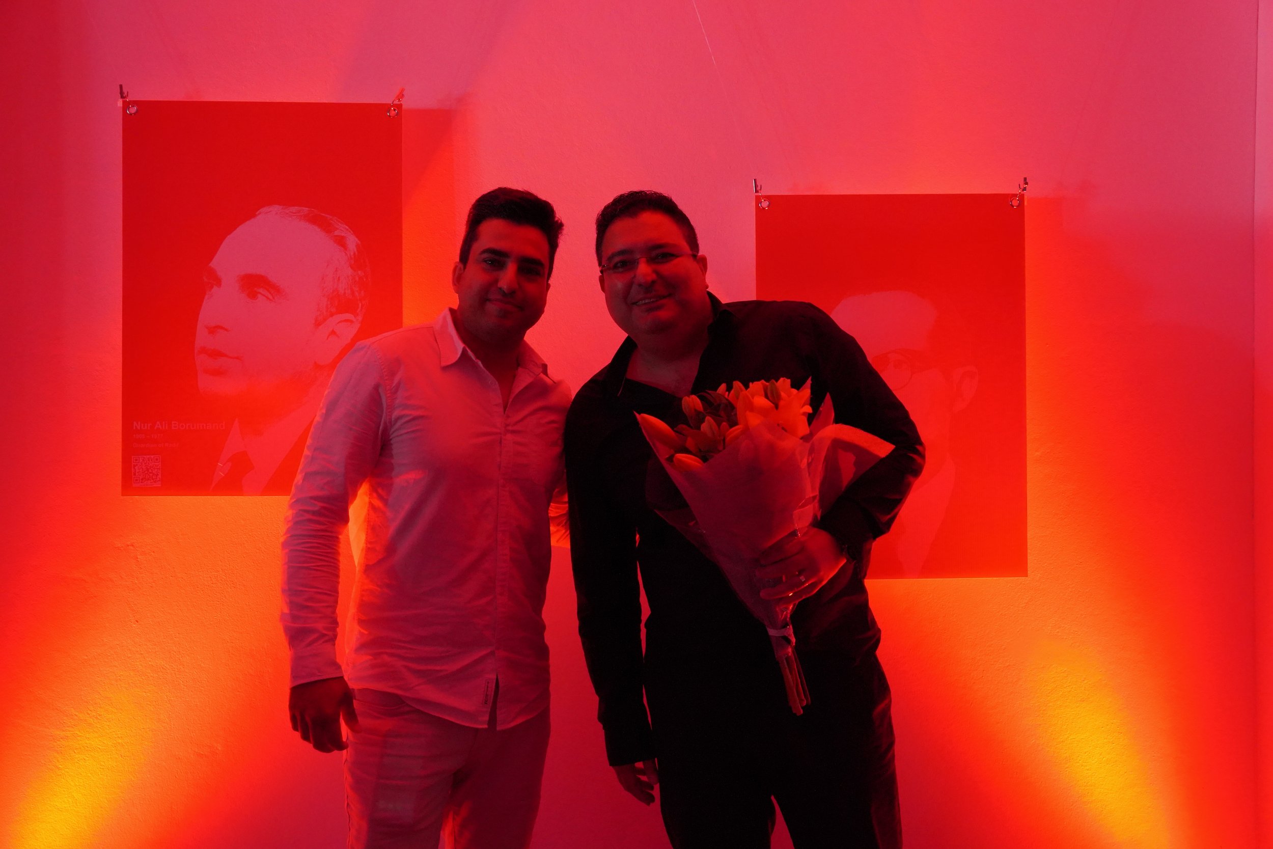 Two men standing in a red-lit room, one holding a bouquet of flowers, with posters of faces on the wall behind them.