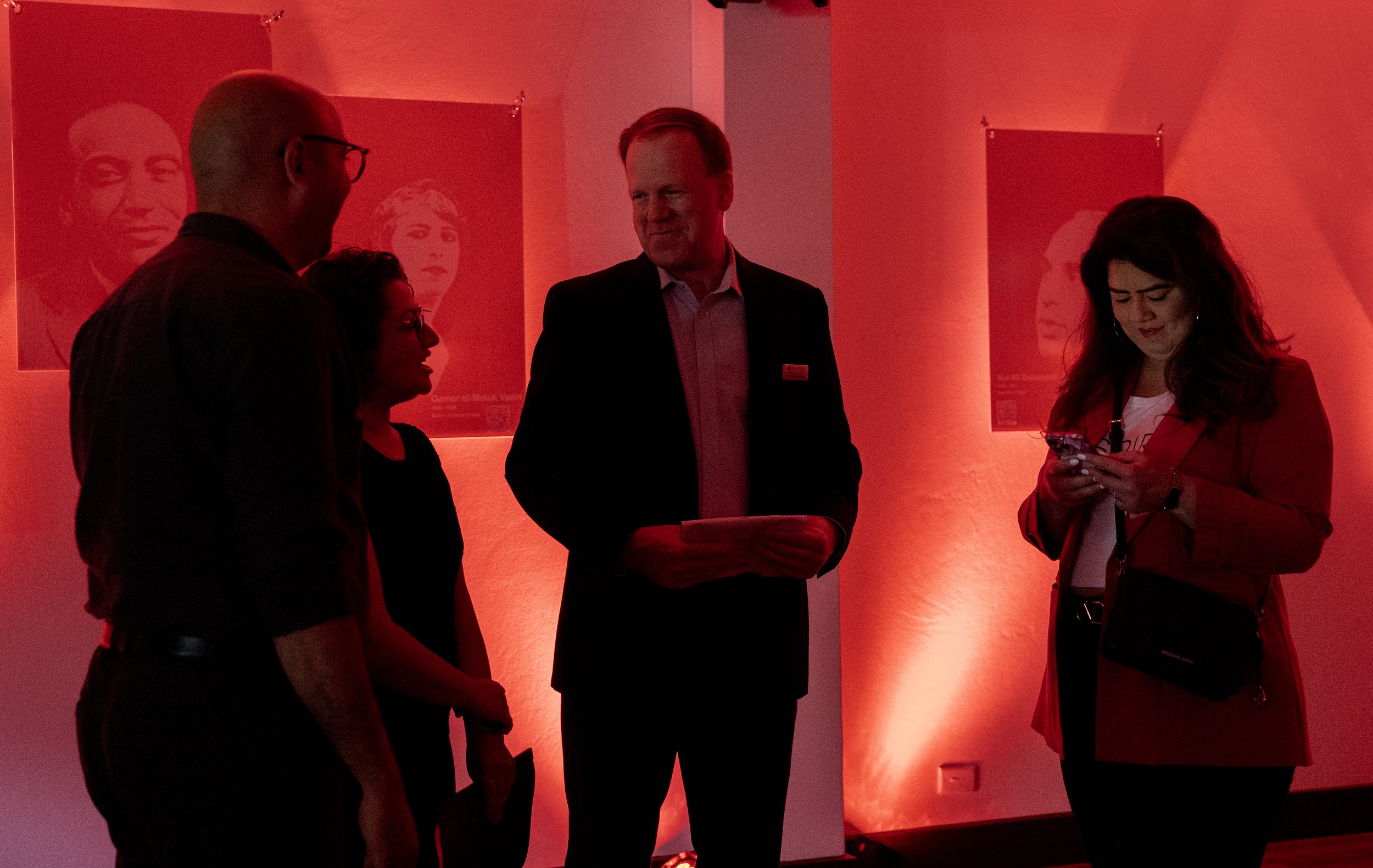 Four people in a red-lit room with portraits on the wall, one person using a mobile phone.