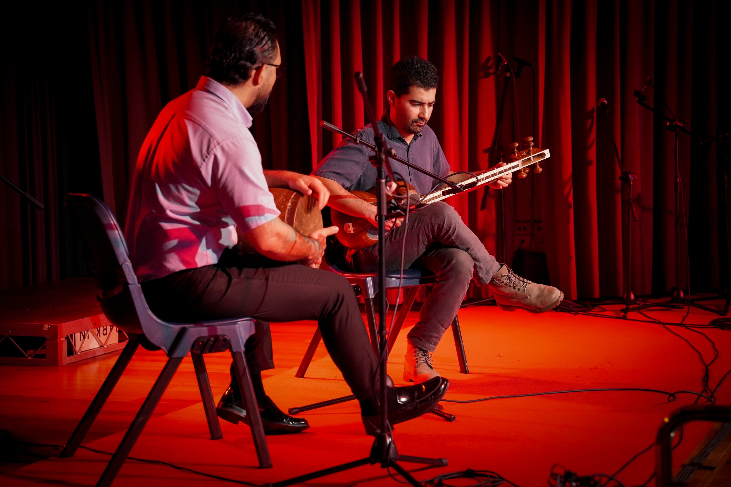 Two musicians perform on stage, one playing a drum, and the other playing a stringed instrument similar to a tar, seated under red lighting and microphones.