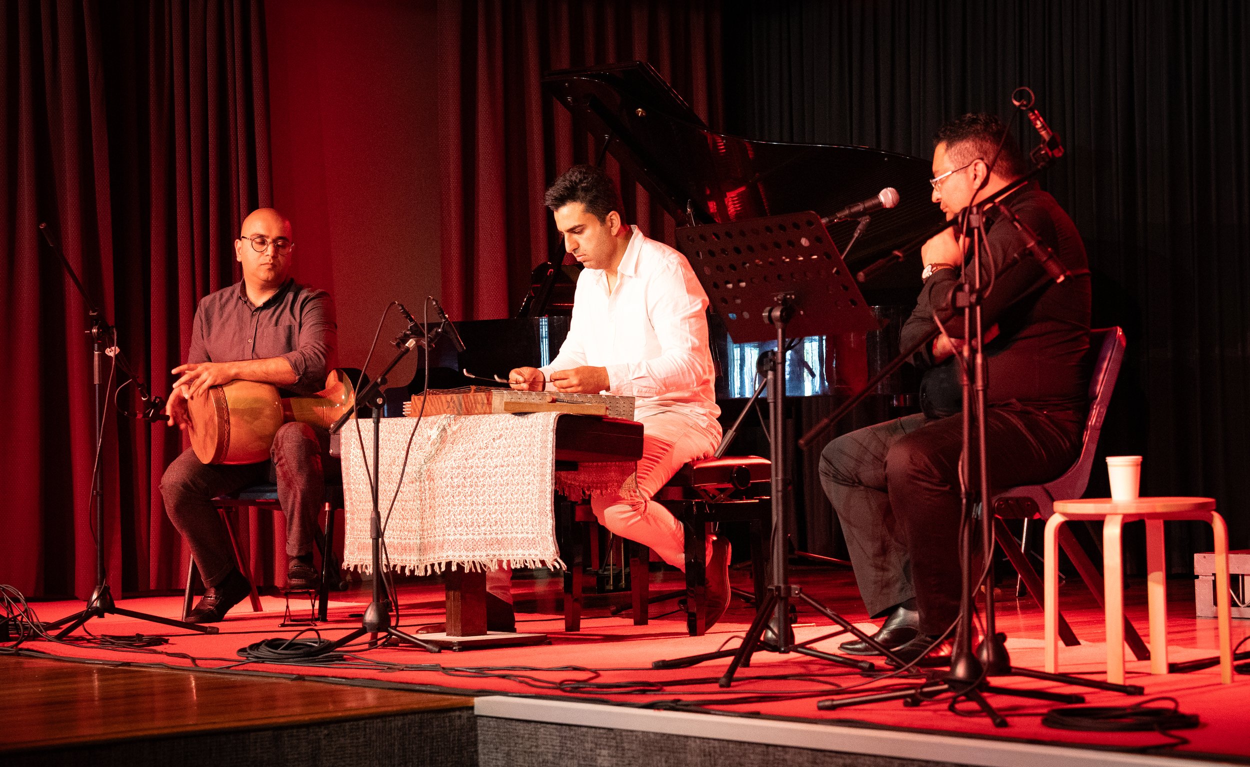 Musicians playing traditional Middle Eastern instruments on stage, including a drum and a string instrument, with microphones and a piano in the background.