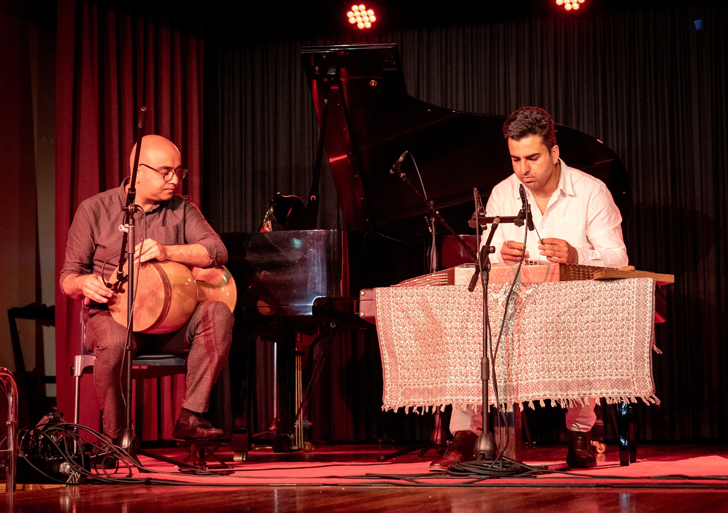 Two musicians performing on stage; one playing a goblet drum and the other playing a hammered dulcimer, with a piano in the background and microphones set up.