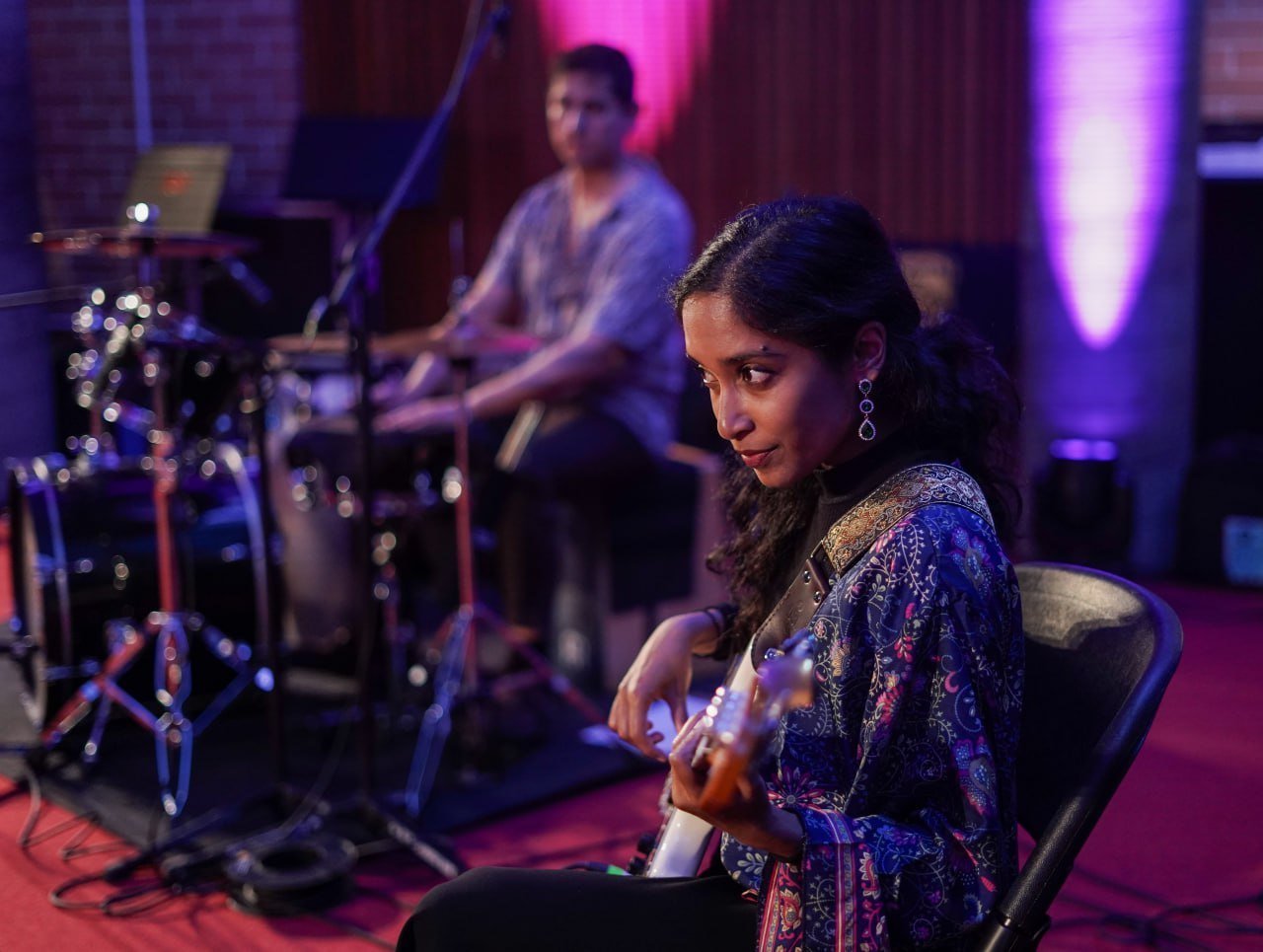 A young woman playing an electric guitar seated in a music studio, with various musical instruments and a person playing drums in the background.