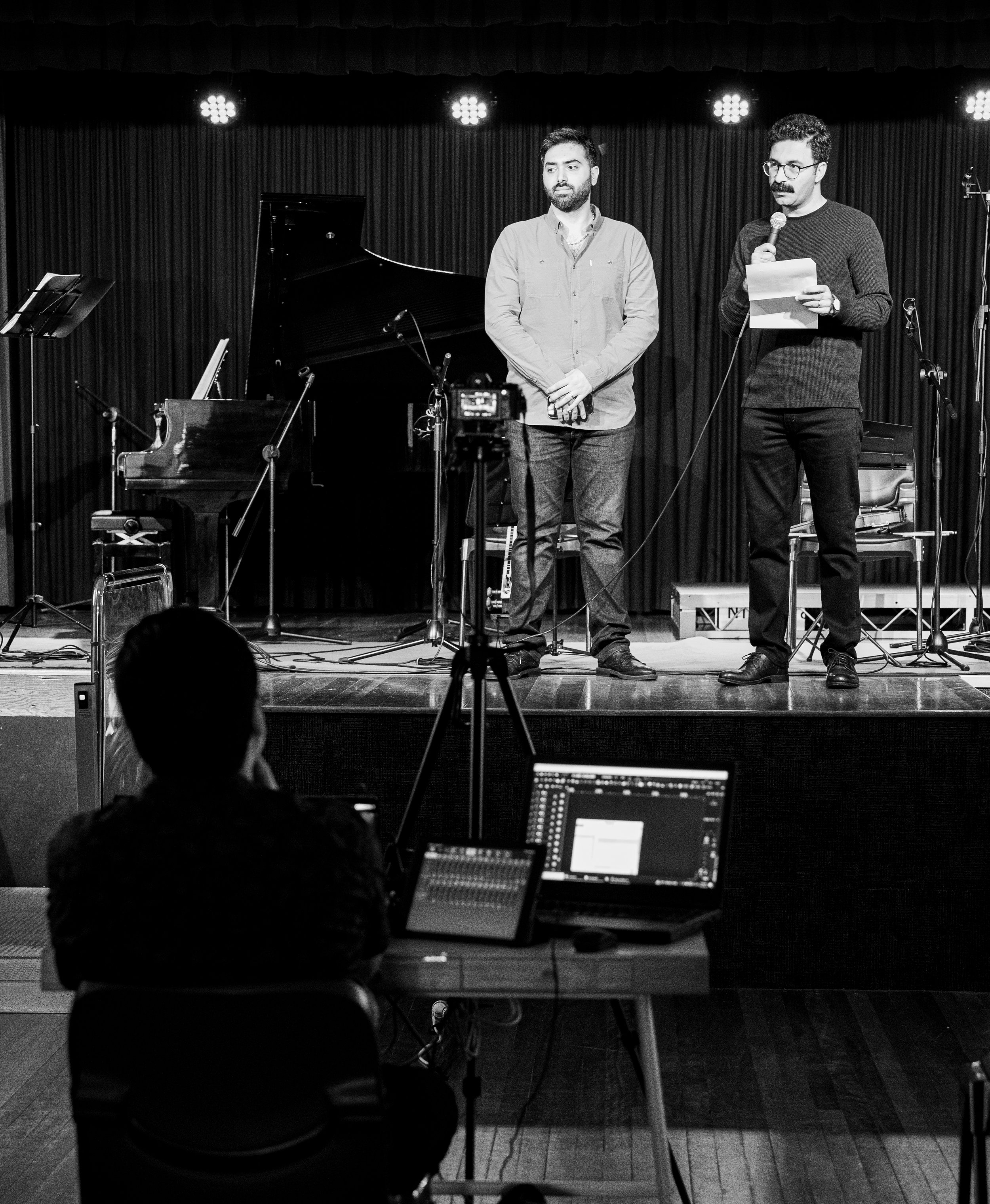 Two men standing on stage with microphones in a performance setting, a grand piano and music stand in the background, and a person at a control station with a laptop in the foreground.