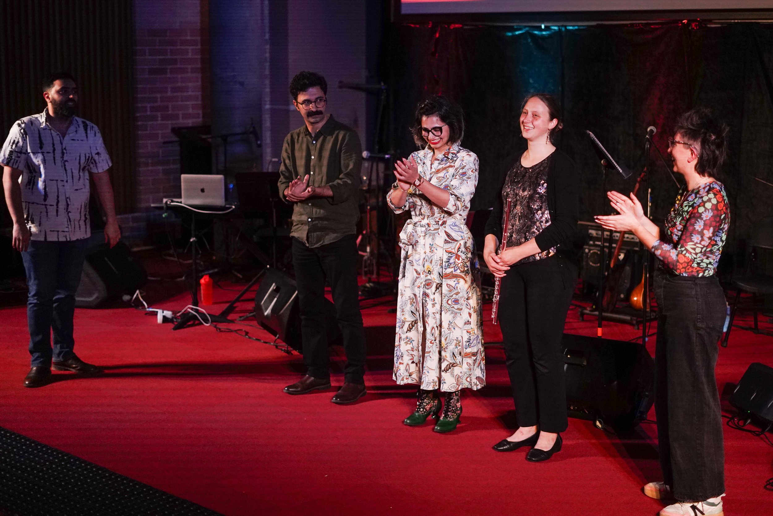 Five people standing on a red carpeted stage, some clapping and one holding a flute, with microphones and musical instruments in the background.