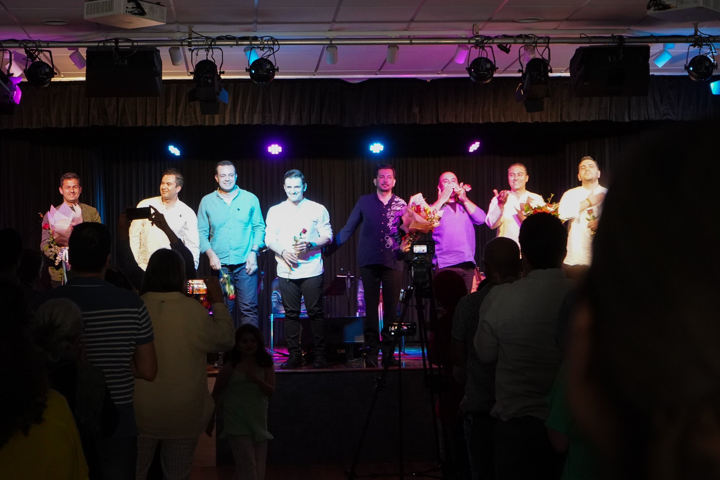 A group of men standing on a stage receiving applause and holding flowers, with an audience in front capturing the moment.