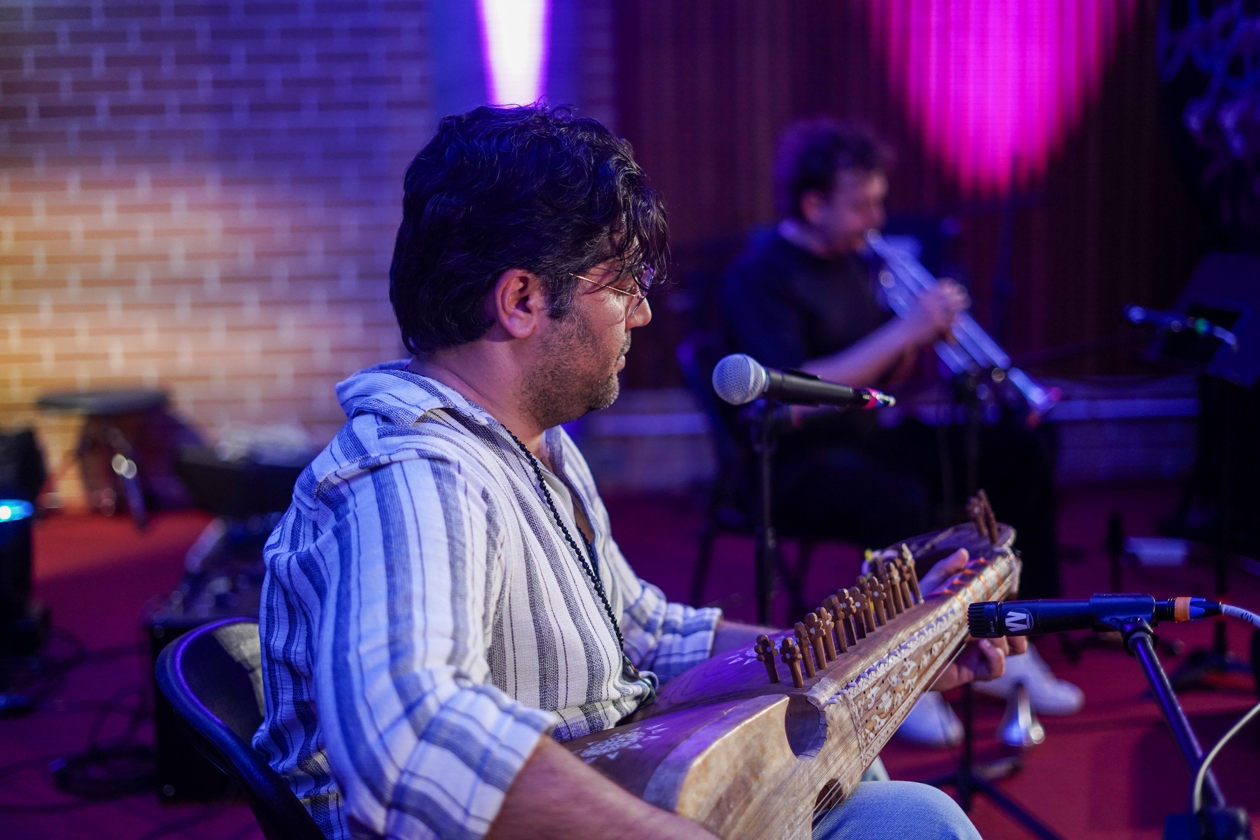 Musician playing a traditional string instrument with a trumpet player in the background, set in a dimly lit performance venue with brick walls.