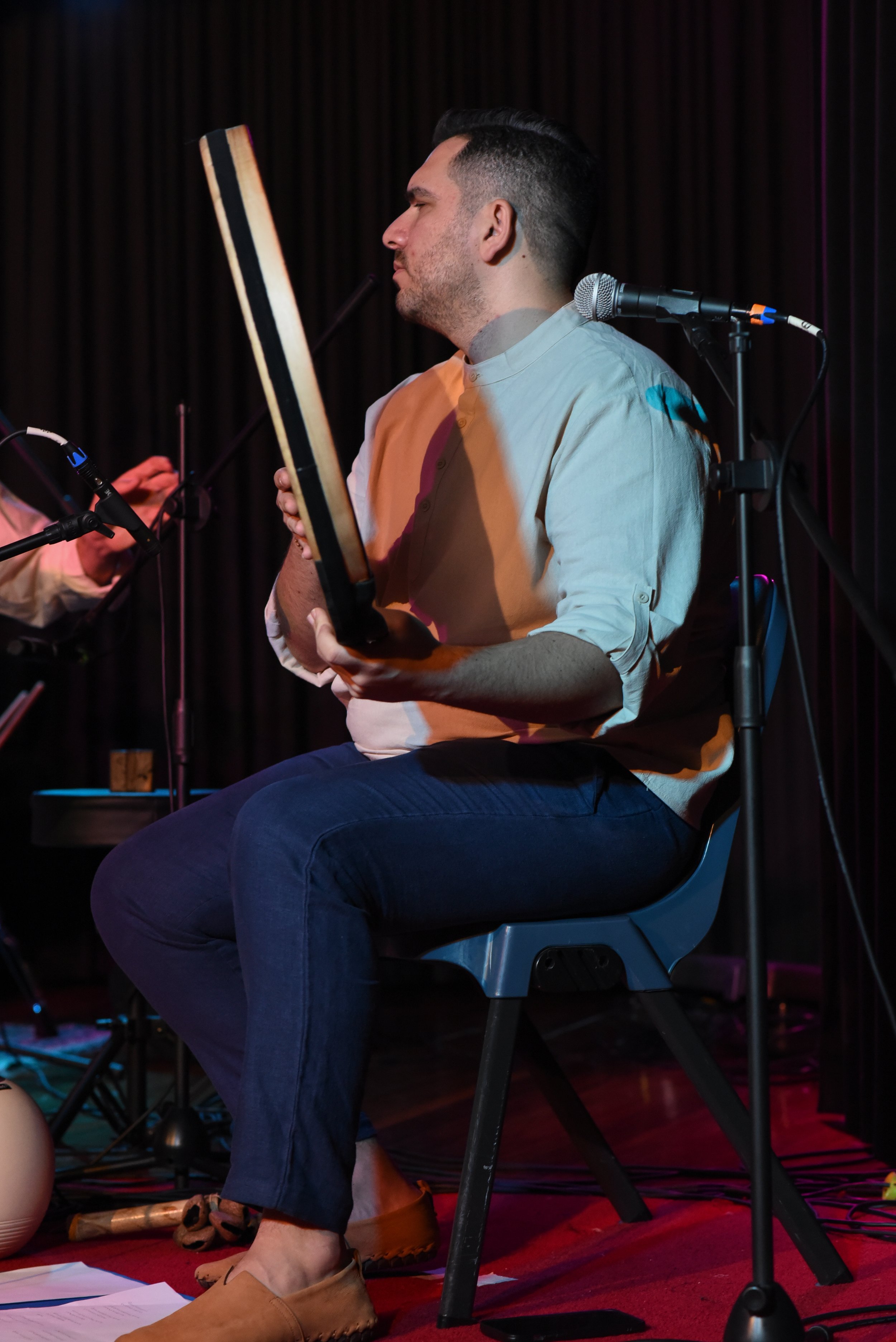 A man sitting on a blue chair playing a drum, with a microphone nearby, on a stage with a red carpet.