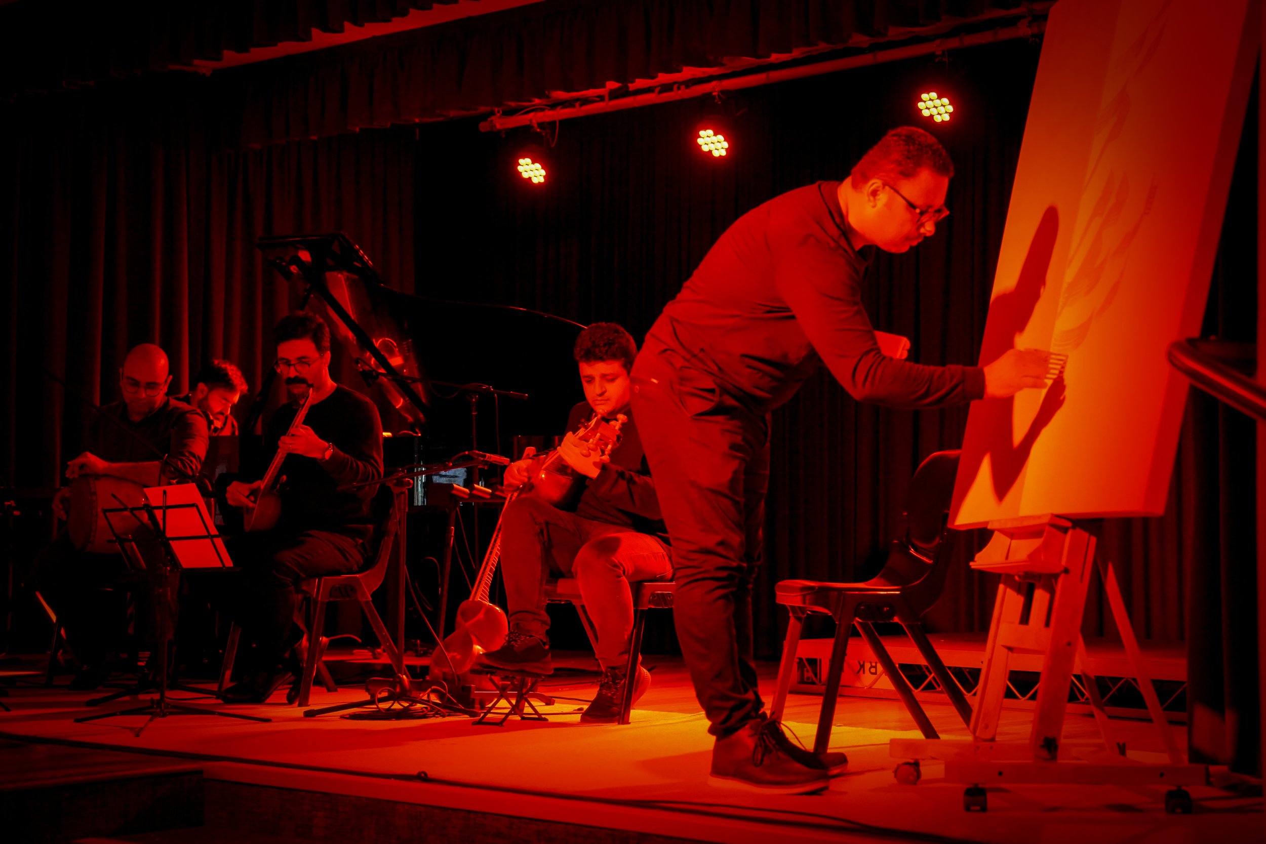 Musicians performing on stage with red lighting while a man paints on a canvas.