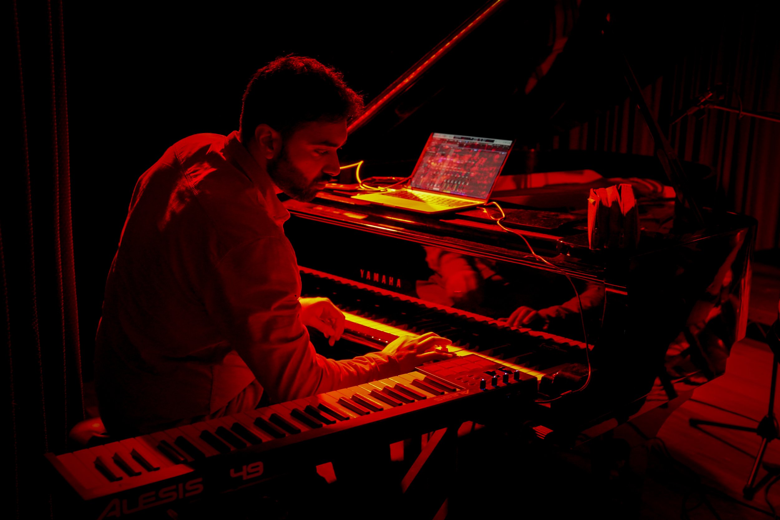 Musician playing piano and keyboard with laptop on piano lid, illuminated by red light, in a dimly lit setting.