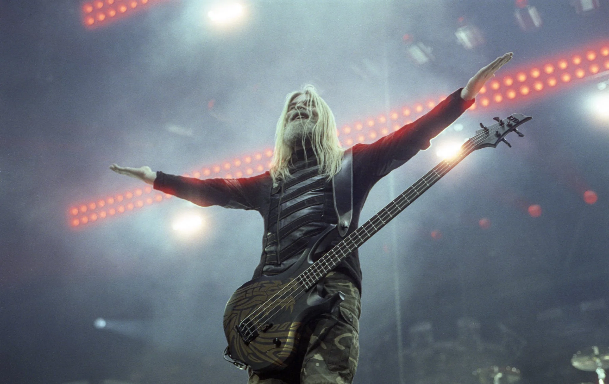 A musician with long blonde hair and a beard, wearing a striped shirt and camo pants, standing on stage with arms outstretched, holding an electric guitar with a tiger stripe design, surrounded by bright stage lights.