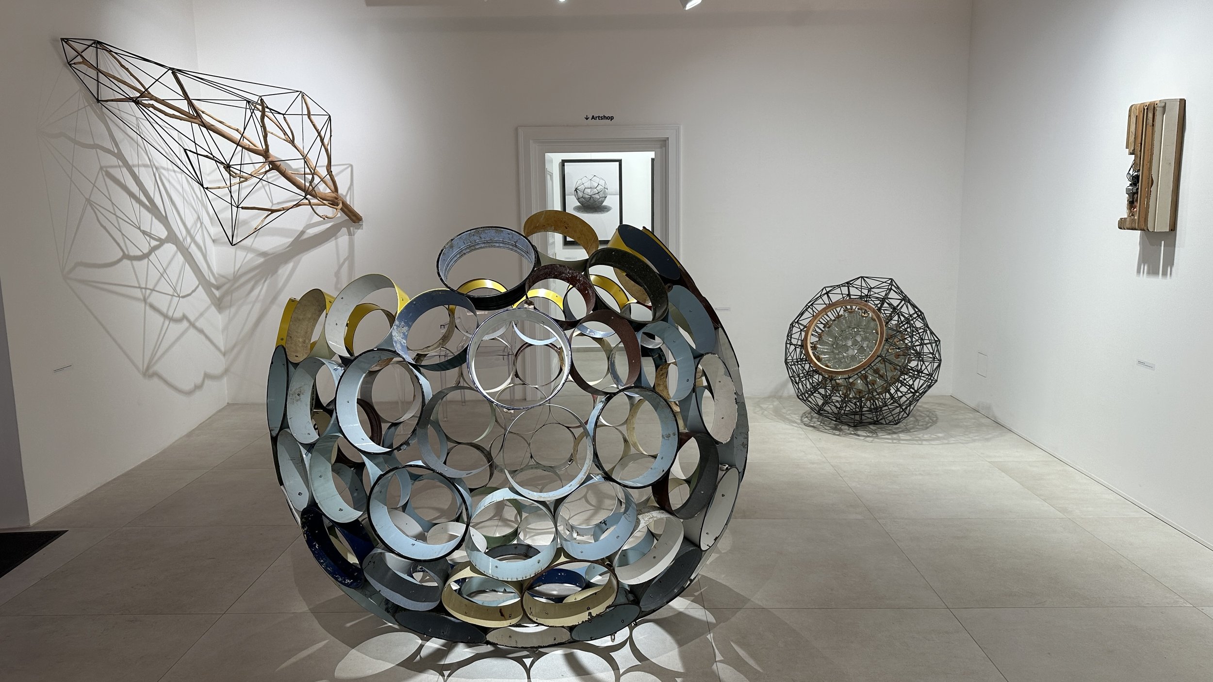 Art installation in a gallery featuring various sculptures. In the foreground, there is a large spherical sculpture made of interconnected metal rings. To the left, a geometric artwork made of wooden sticks and metal wires casts shadows on the wall. 