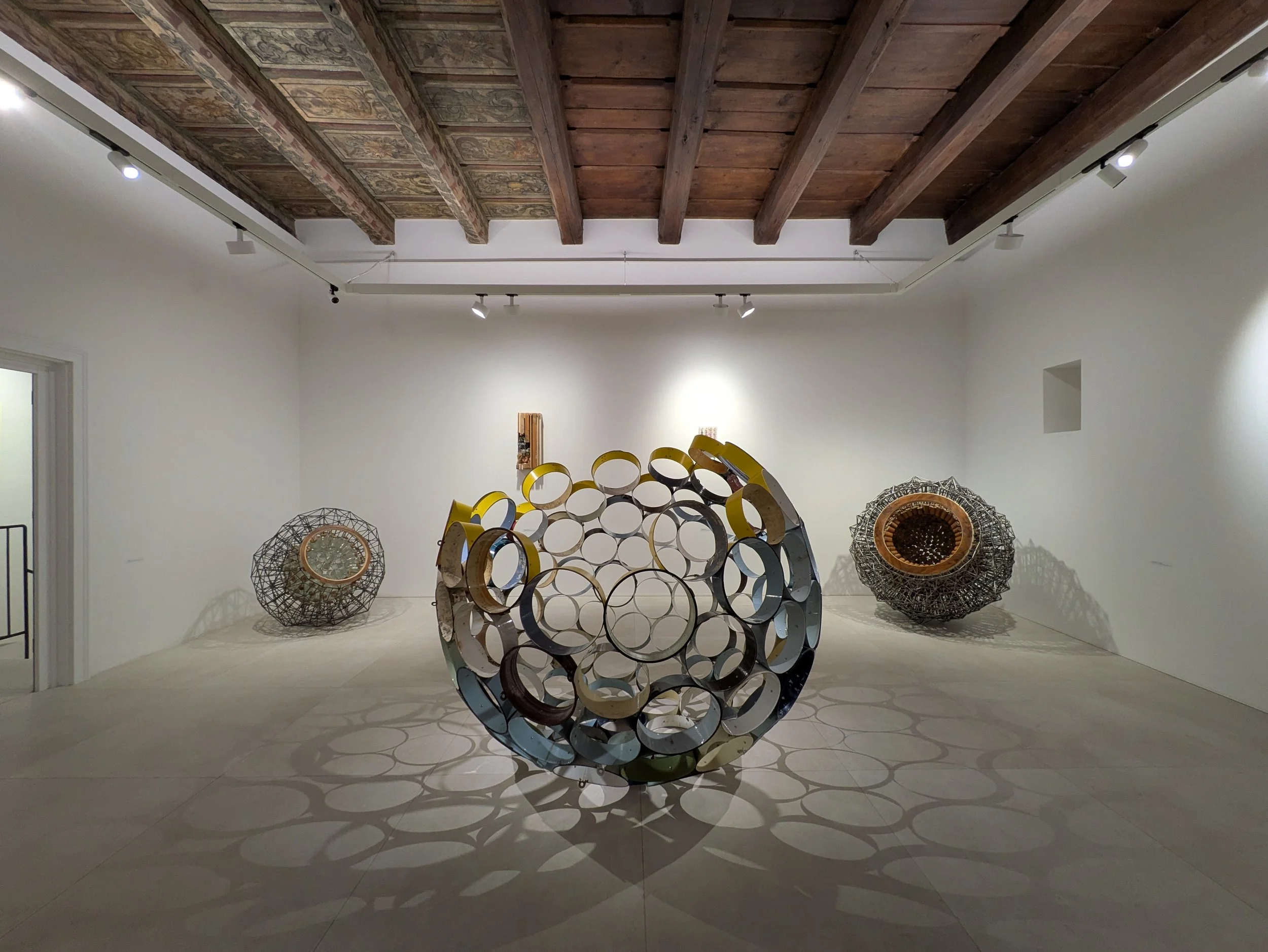 Art gallery with three spherical sculptures made of circular rings, casting shadows on the floor, with a wooden ceiling and white walls.