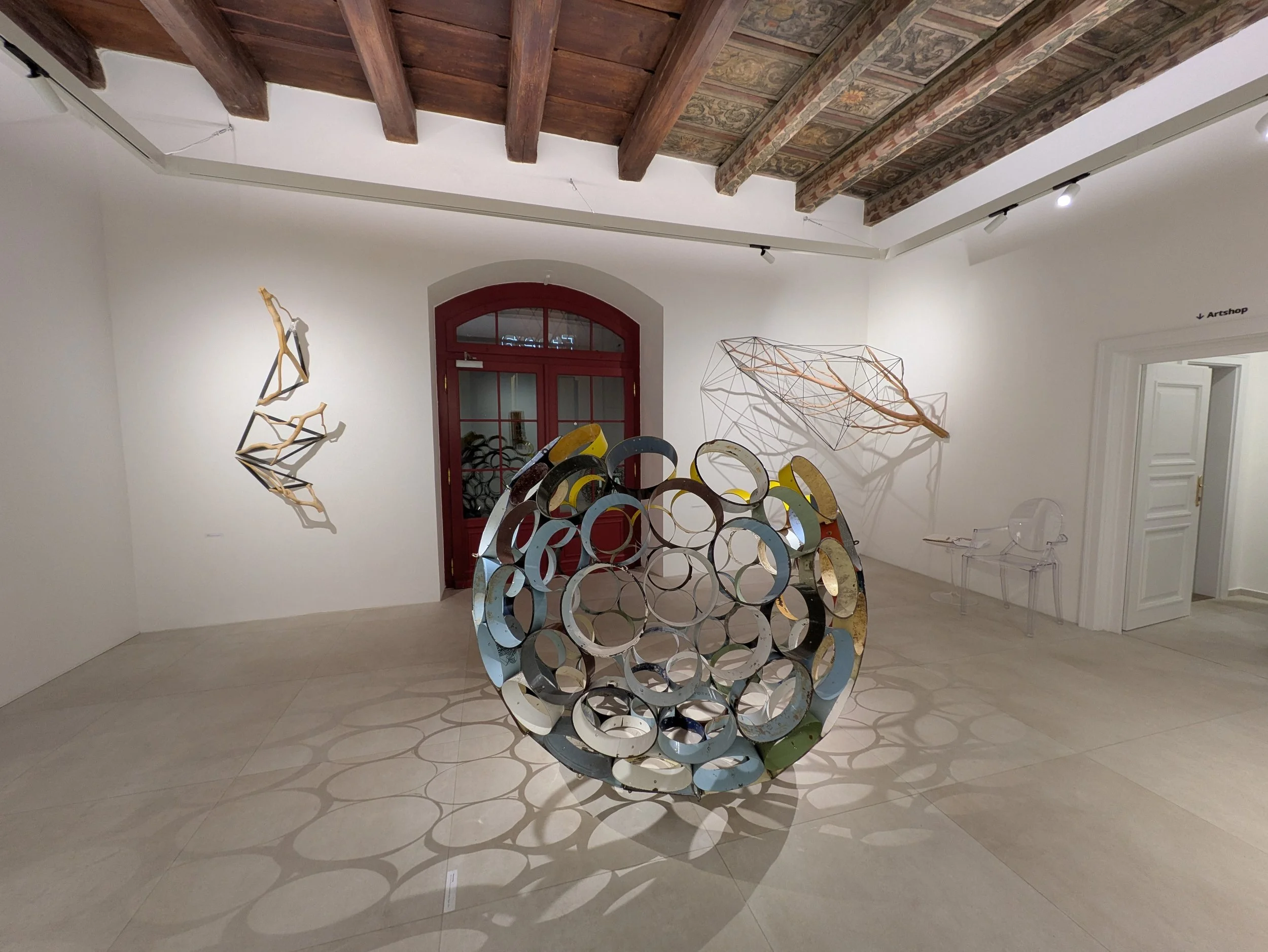 Contemporary art gallery featuring a spherical sculpture made of interconnected circular metal rings in the center, with abstract wall sculptures of wooden and metal geometric shapes and shadows on white walls, wood-beamed ceiling, and a curved red d