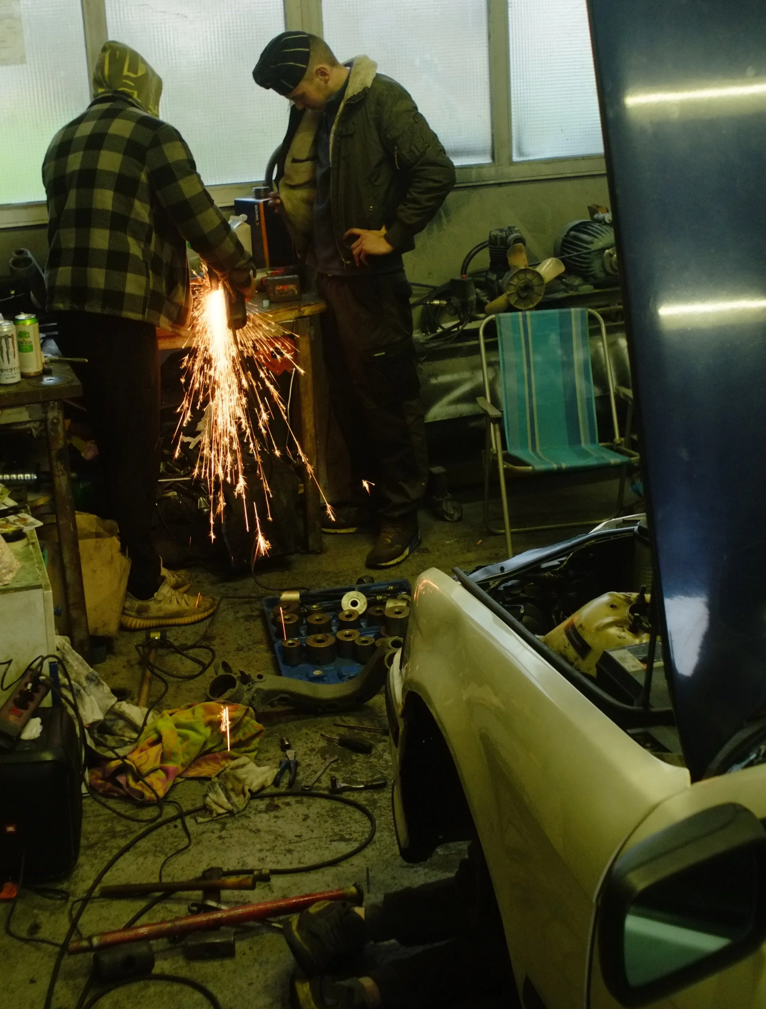 Two people in a garage working on a car with one using a metal grinder, producing sparks, and various tools and parts scattered around.