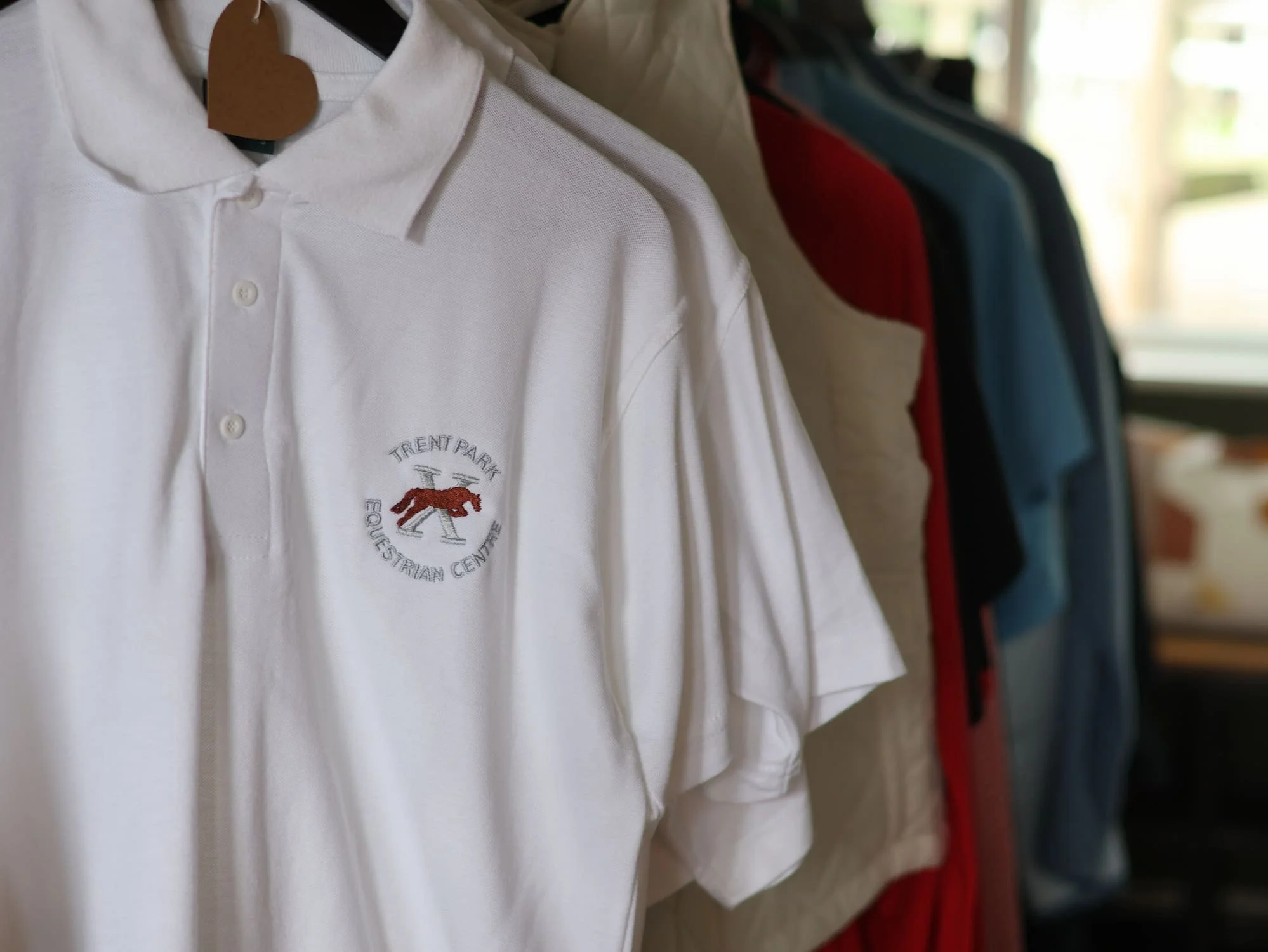 Close-up of a white polo shirt with embroidery on the chest area, hanging on a rack with other shirts in various colors in the background.