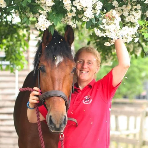 A woman with short blonde hair in a red shirt smiling next to a brown horse, holding a bunch of white flowers above them in an outdoor setting.