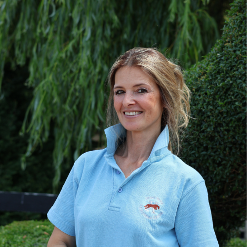 Smiling woman in a light blue polo shirt standing outdoors with green trees and bushes in the background.