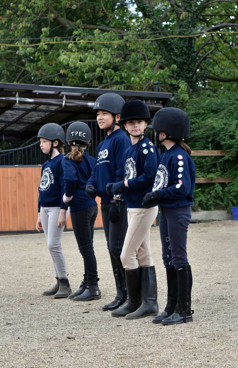 Five young girls wearing helmets, navy blue sweatshirts, and riding boots are standing in a line outdoors. They appear to be at a riding or equestrian camp, as indicated by the emblem on their sweatshirts.