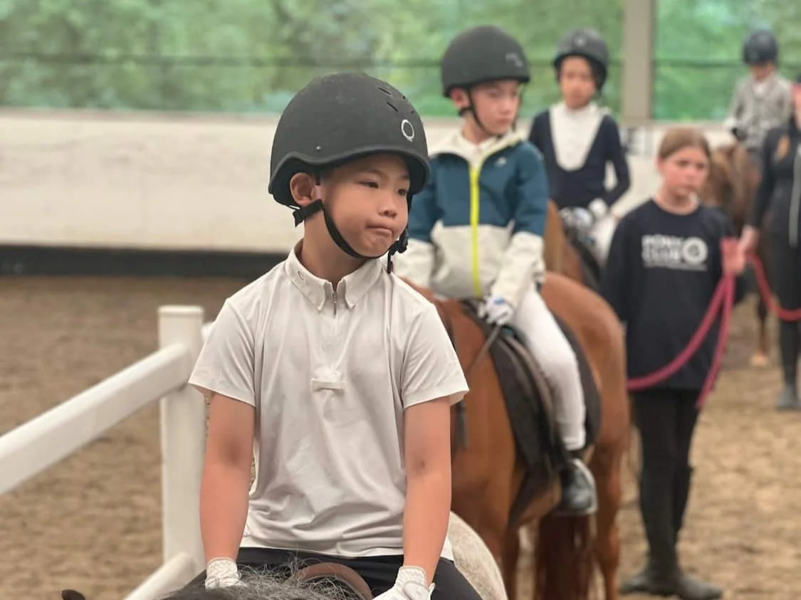 Children wearing helmets riding horses in an indoor riding arena.