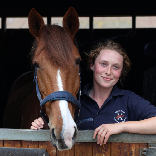 A young woman and a chestnut horse with a white blaze on its face inside a stable.