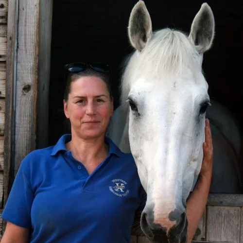 A woman in a blue polo shirt standing next to a white horse, both facing the camera. The woman has dark hair and is outdoors near a wooden building.