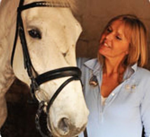 A woman with shoulder-length hair and a light blue shirt looking at a white horse with a black bridle in a stable or barn.